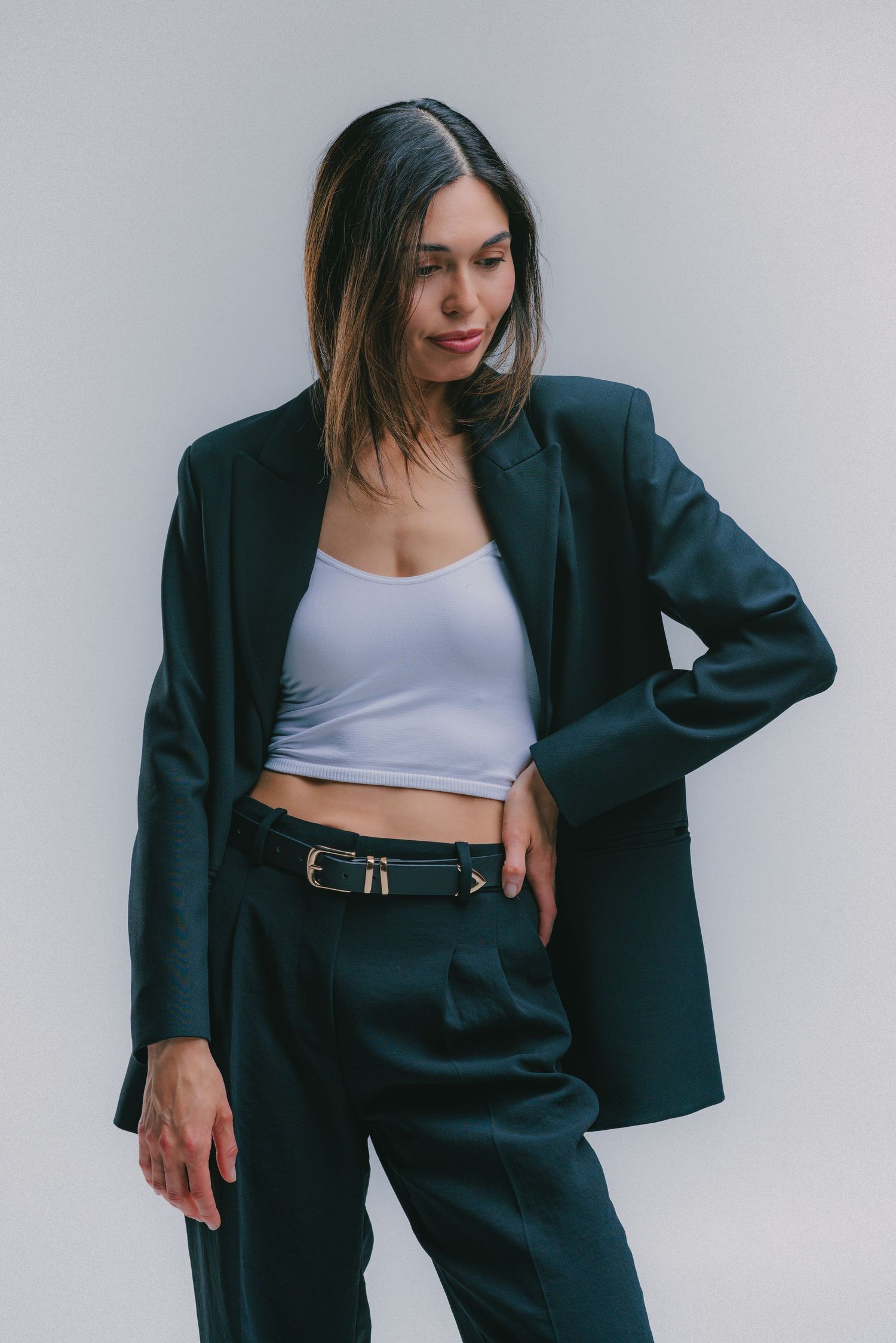 Woman in dark green blazer and trousers with white crop top and belt, posing against light background.