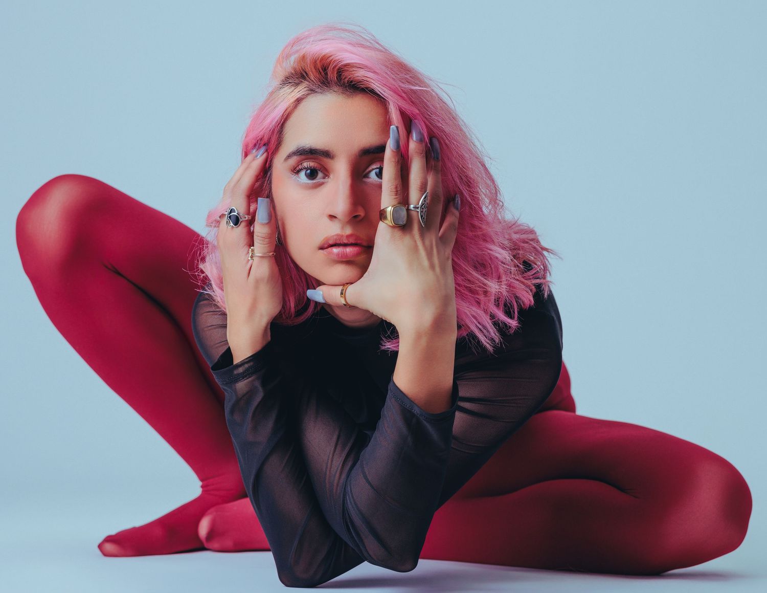 Woman with pink hair wearing red tights and black top, posing on floor with rings on fingers.