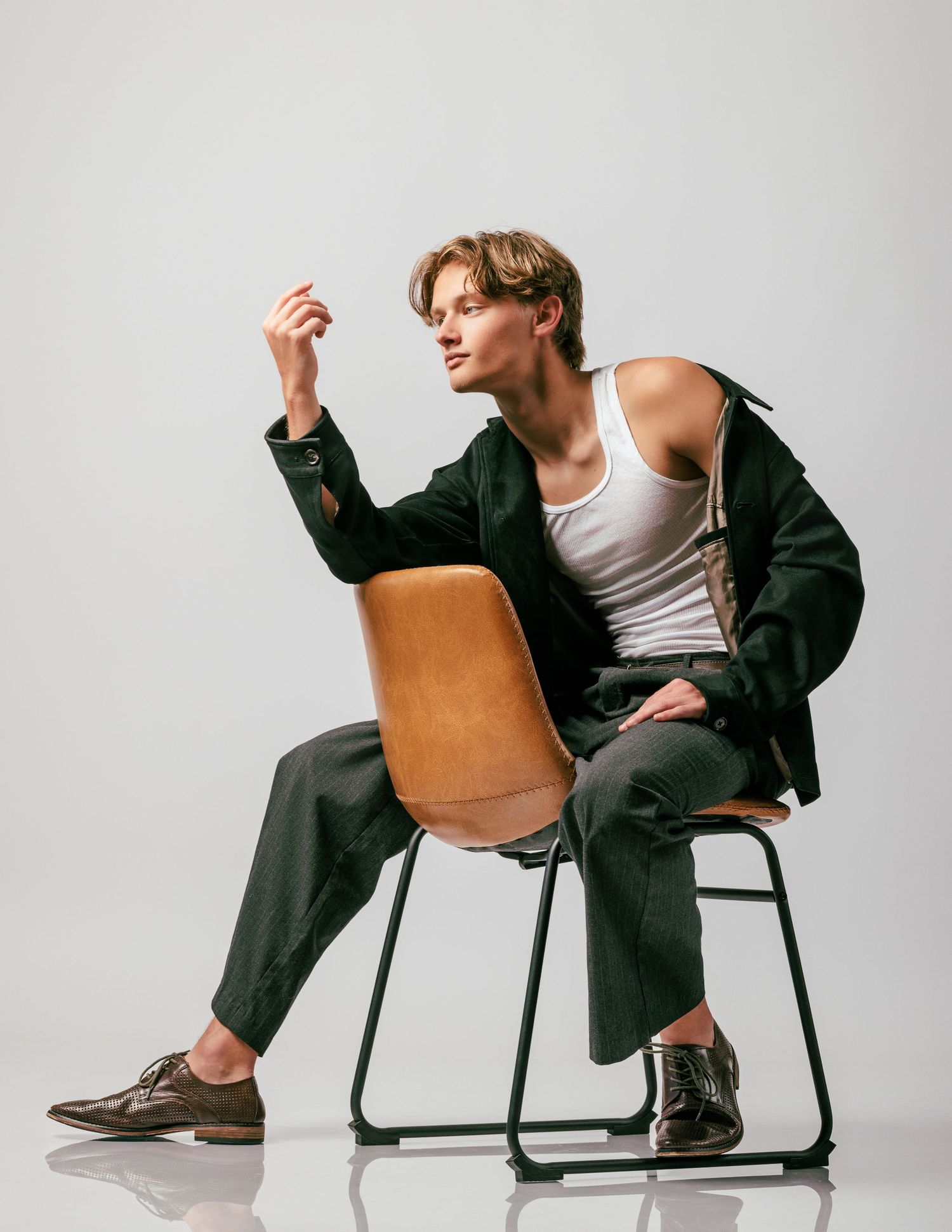 Young man in dark jacket and white tank top seated on wooden chair, posing confidently against white background.