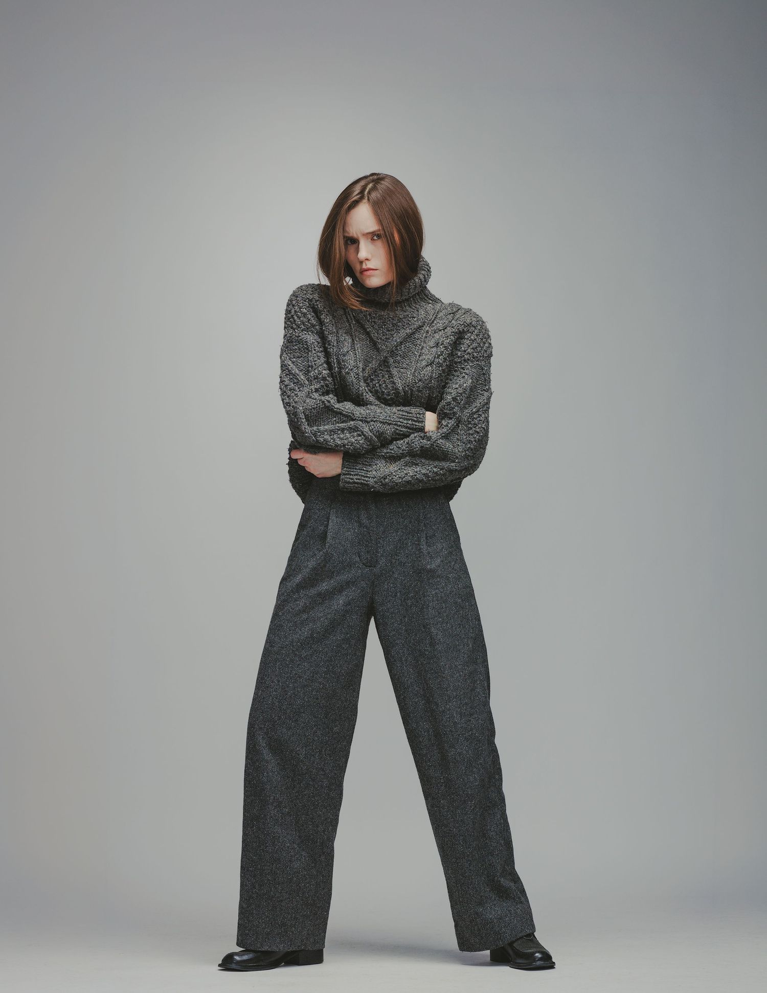 Woman in gray cable-knit turtleneck sweater and wide-leg trousers, arms crossed, posing against neutral background.
