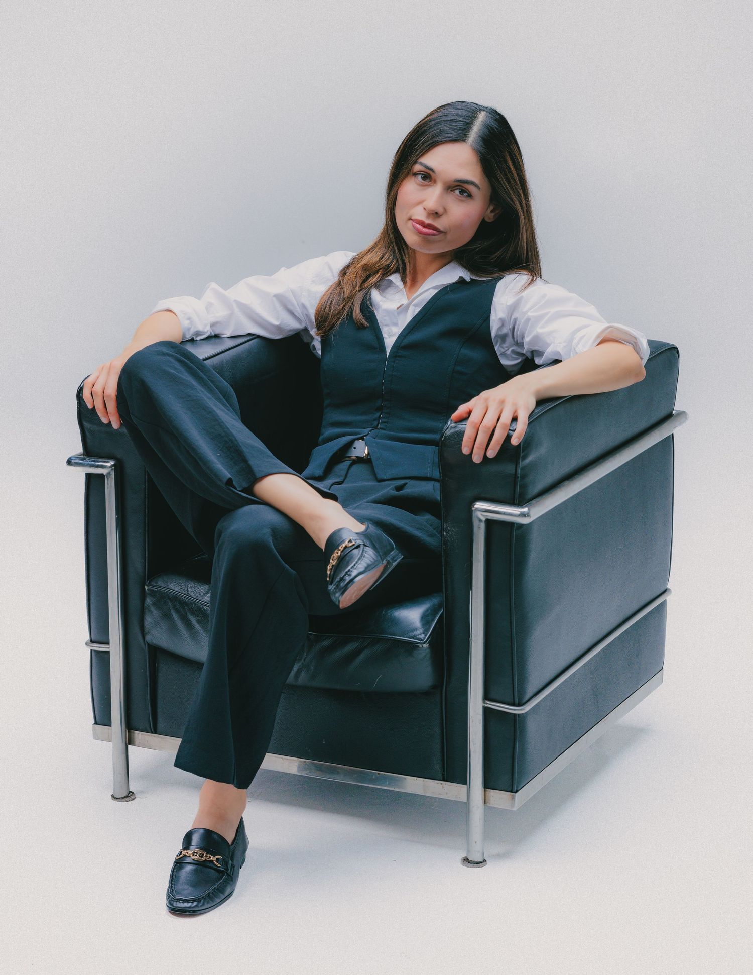 Woman in navy vest and trousers seated confidently in a black leather Le Corbusier chair against a white background.