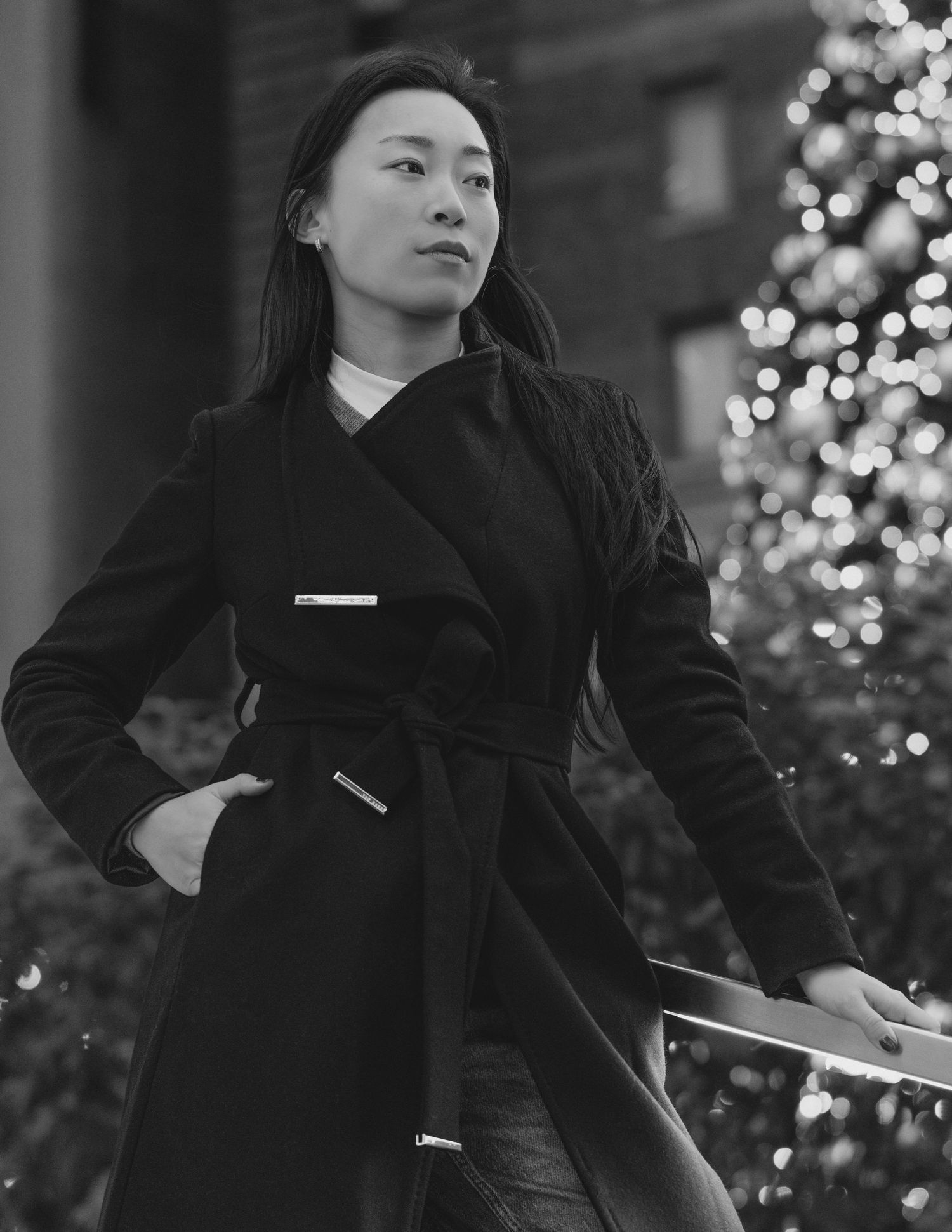 A person in a black outfit poses against a backdrop of twinkling holiday lights in an artistic black and white photo series.