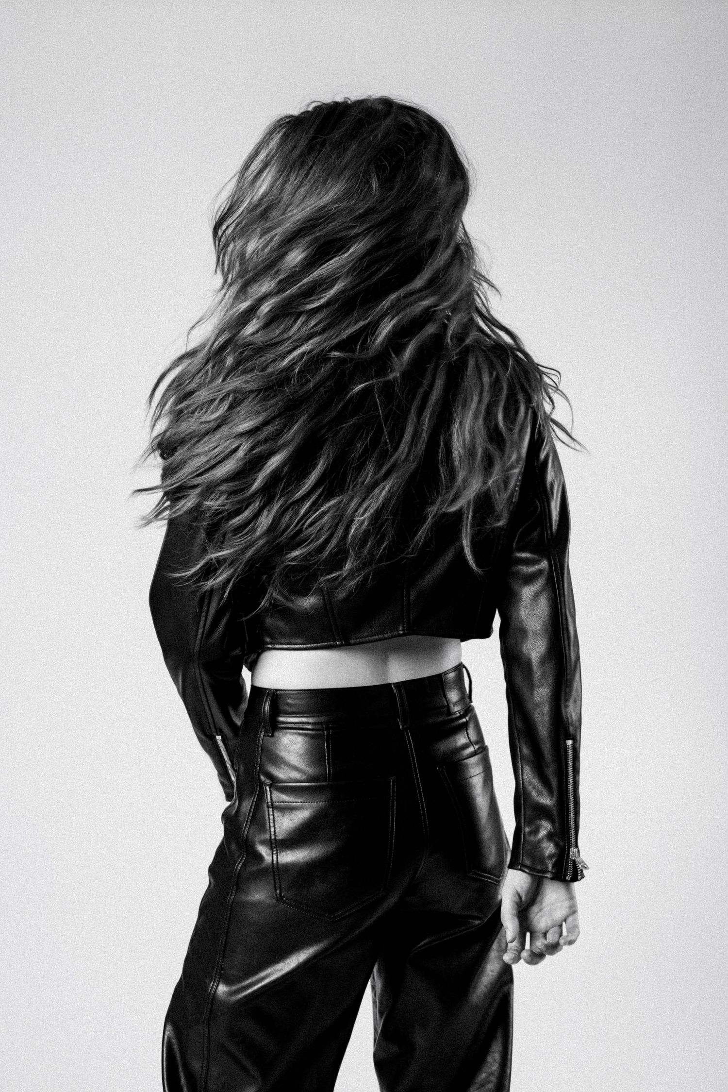 Black and white fashion photo showing person in leather jacket and pants with long flowing hair turned away from camera.