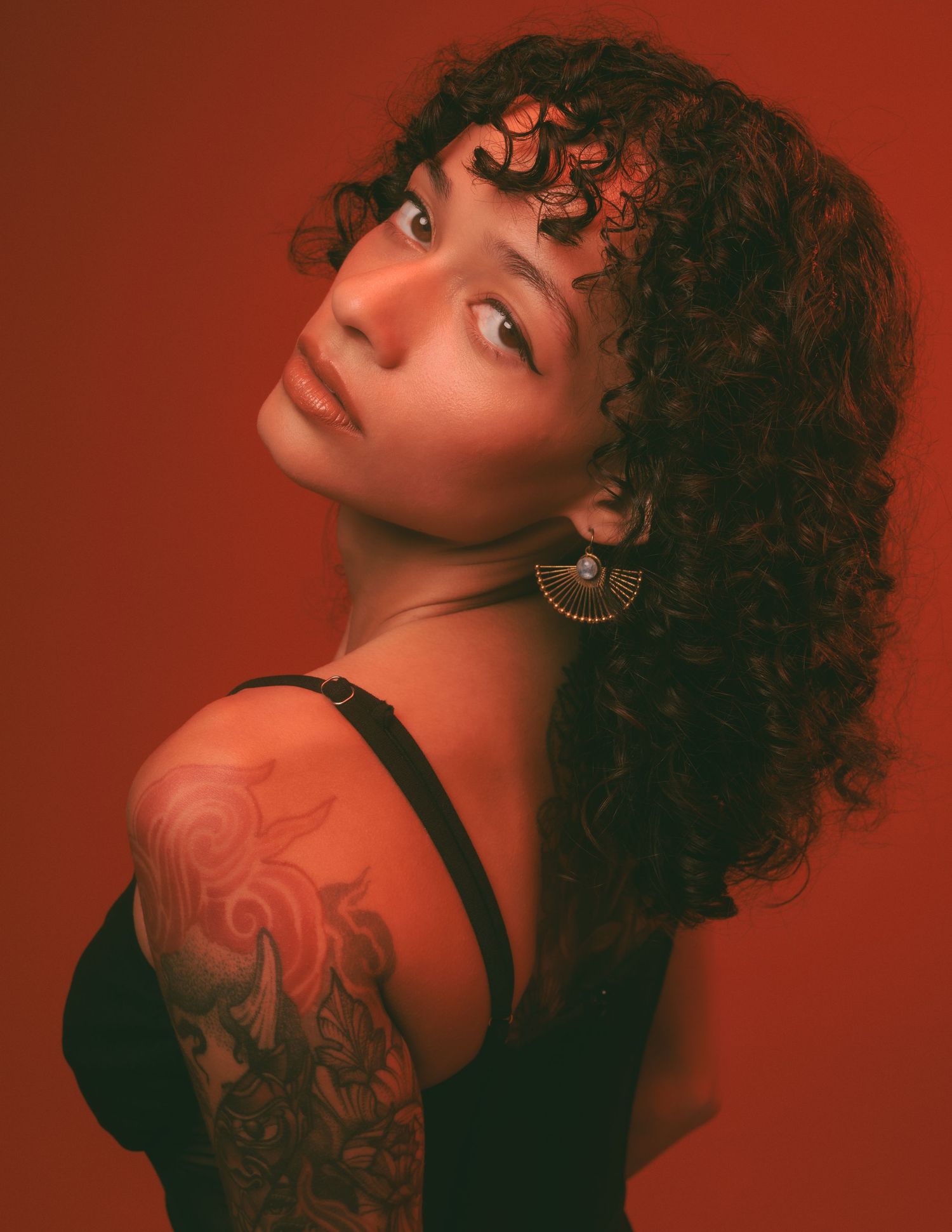 Woman with curly dark hair and a fan-shaped earring looks over her tattooed shoulder in a red-toned studio portrait.