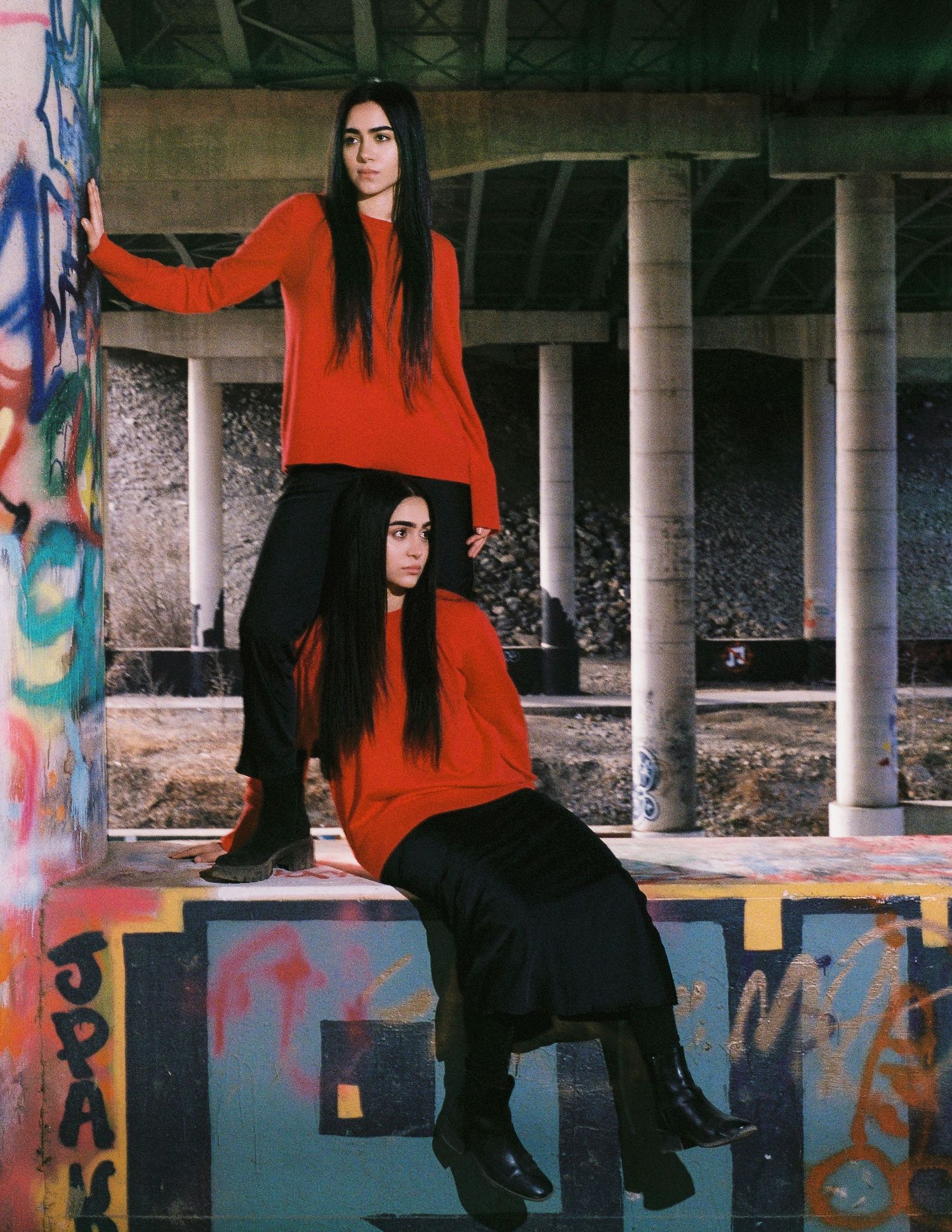 Two people in matching red sweaters and black pants pose artistically in an urban setting with graffiti and concrete pillars.