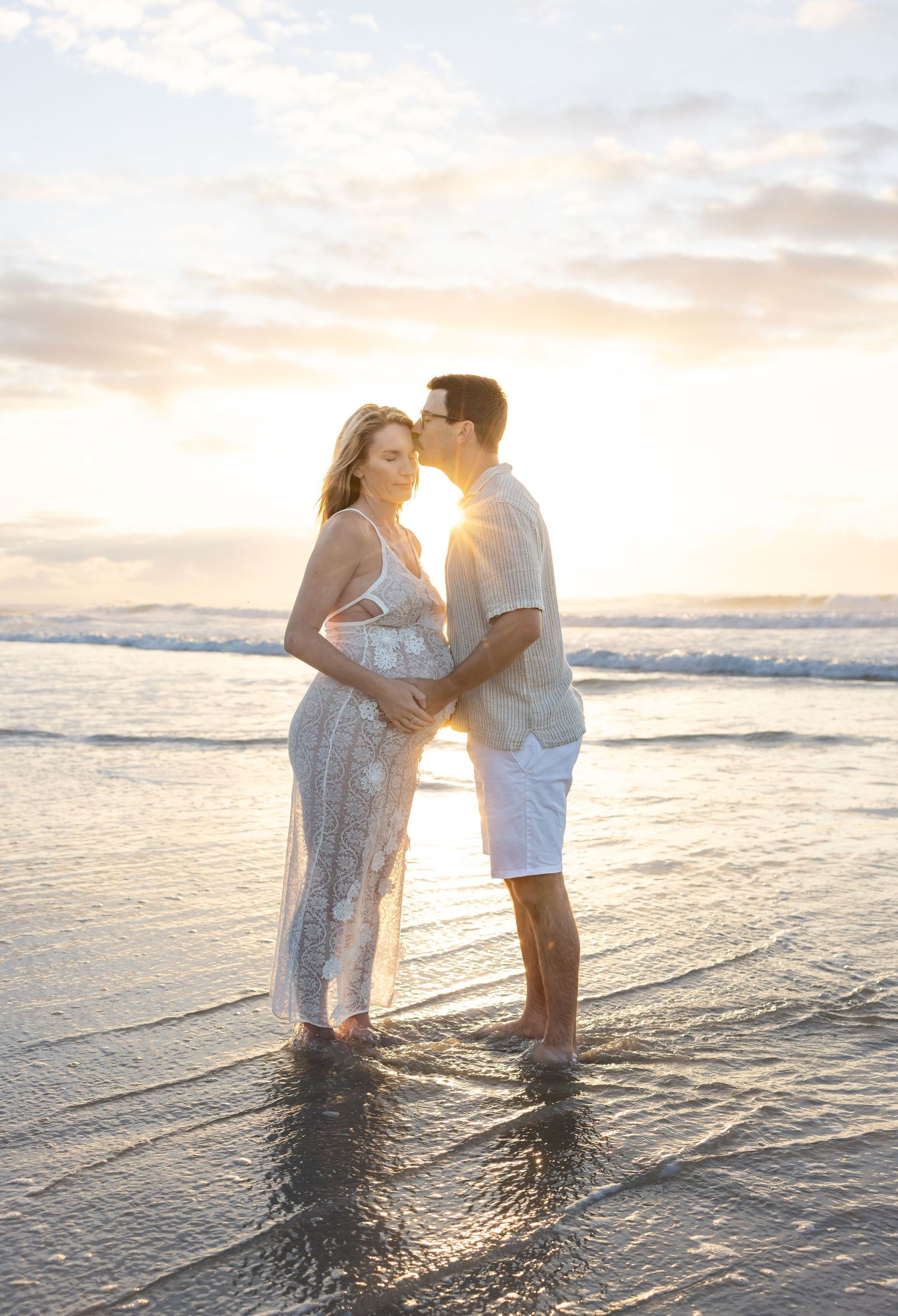 Expecting couple stand holding their unborn child with the sun glowing through the middle of them.