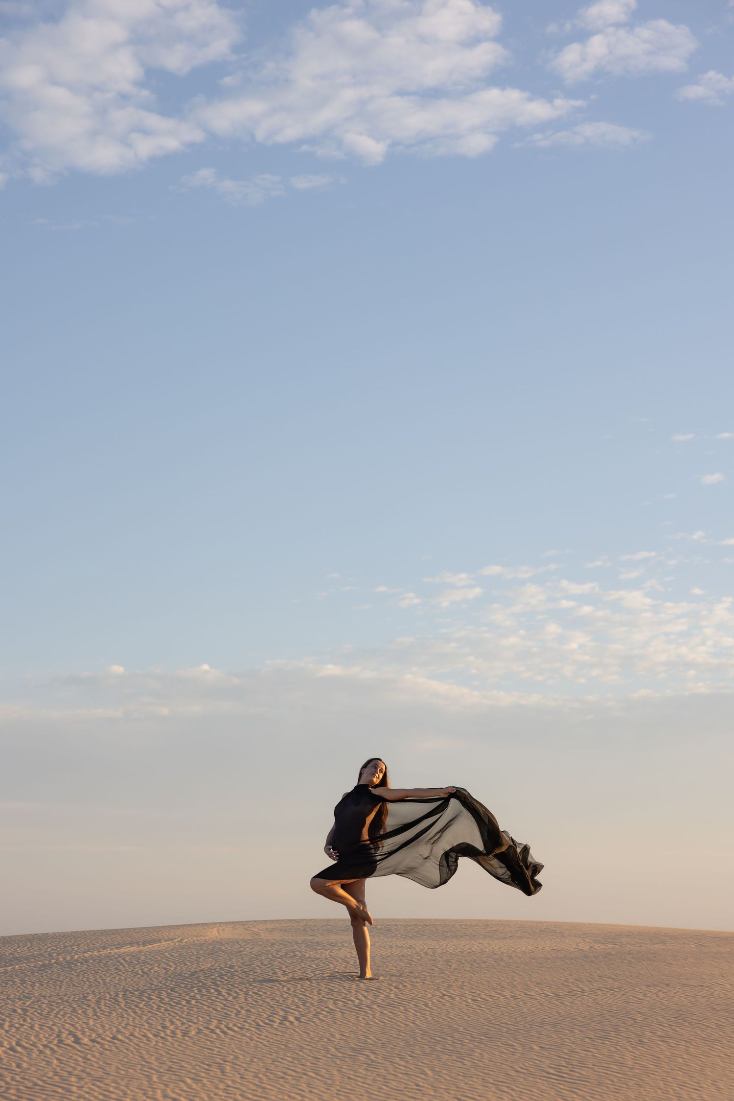 Pregnant mum with flowing black dress stands on the sand with the blue of the sky reaching high above her head.
