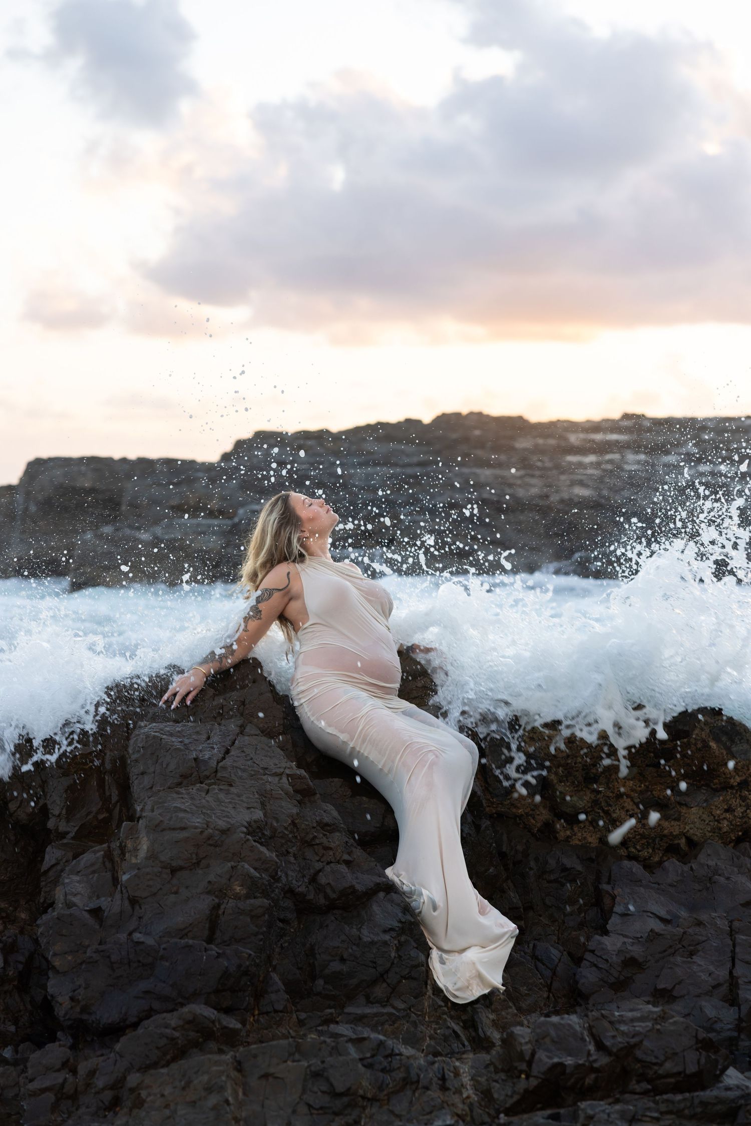 Pregnant mother sits on rocks as gentle waves crash around her at the beach.