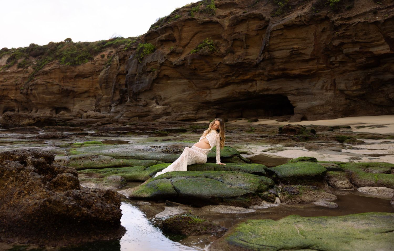 Pregnant mum sitting on the moss covered rocks at Caves Beach, with reflection off the water around the rocks.