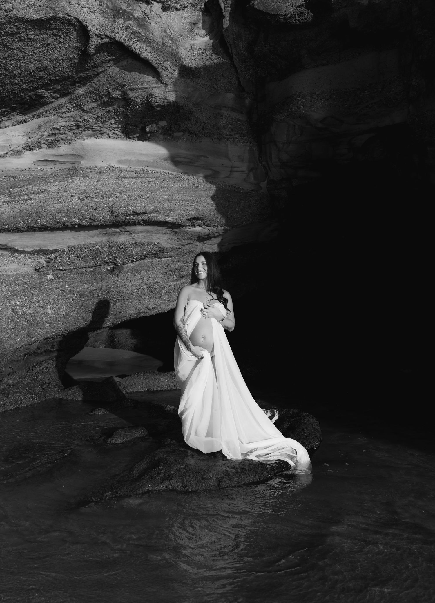 Maternity mum frames her belly with white material, shadow and rocks behind her.