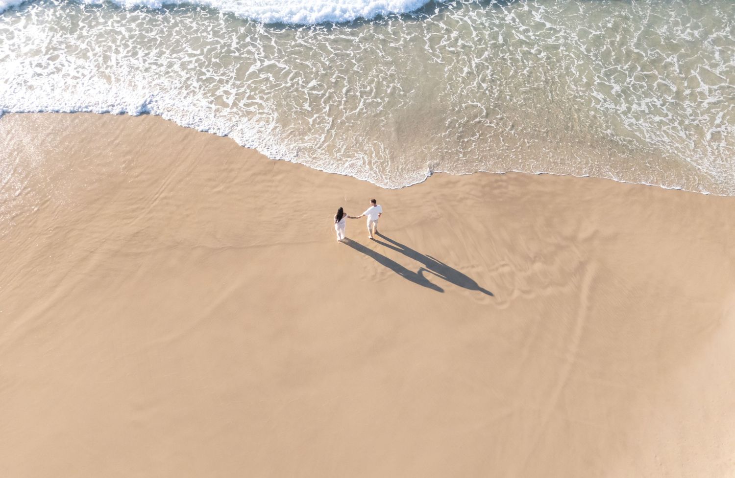 Aerial view of a pristine sandy beach with turquoise ocean waves rolling onto the shore and long shadows cast on the sand.