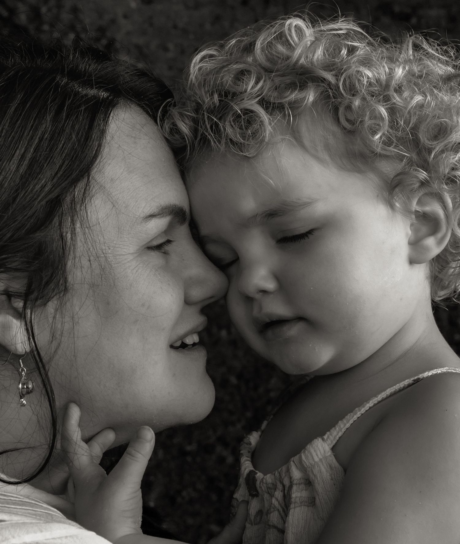 Sleepy child gently rests her face against her mother, touching her mums cheek and closing her eyes. Mother smiles with wonder.
