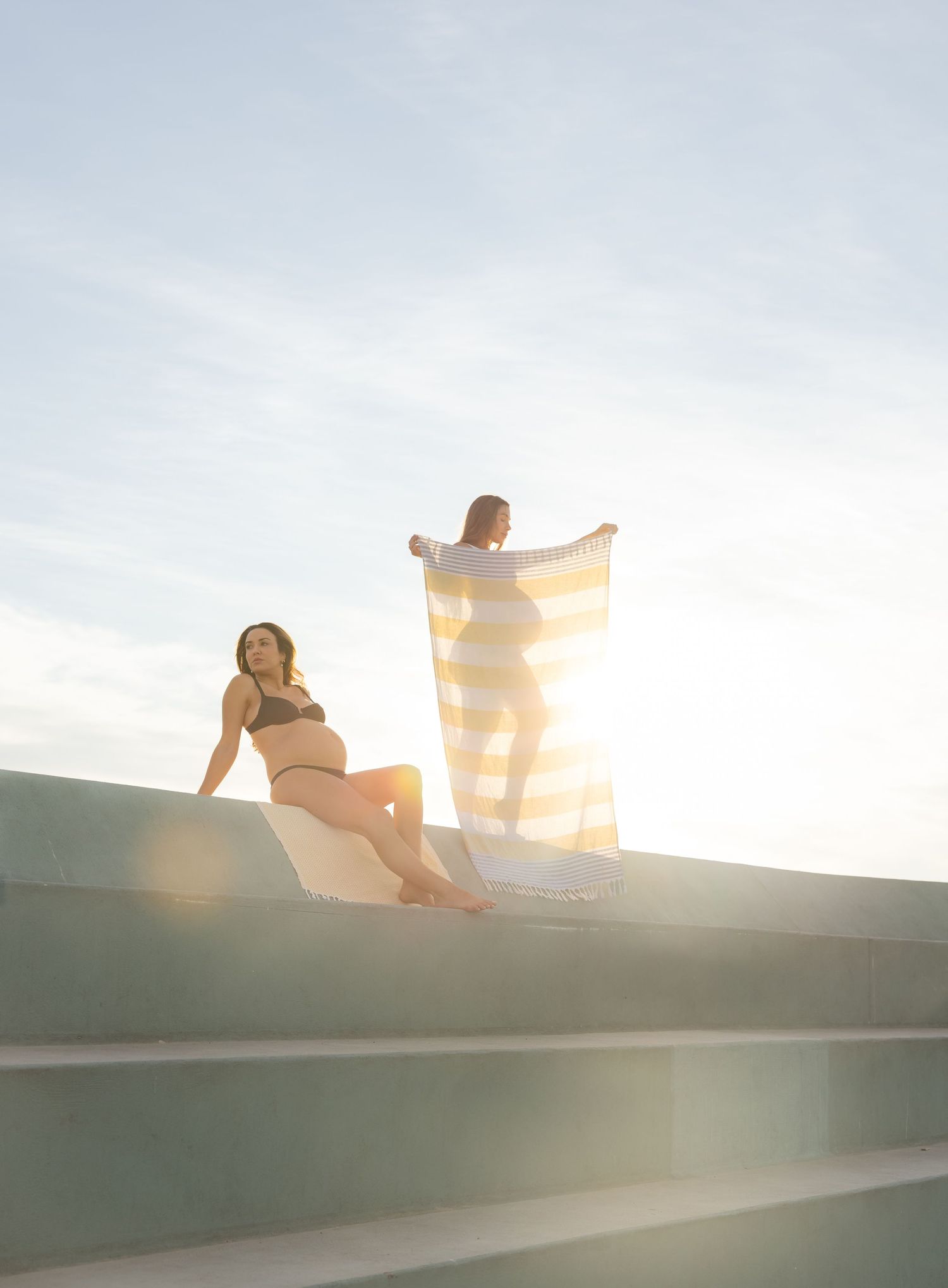 Pregnant friends having a day at Newcastle Baths. One sits looking over the steps and the other is getting ready to lay out a beach towel.