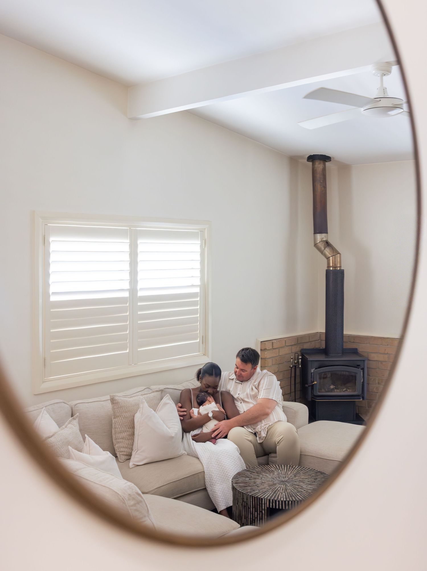 Husband and wife cradle their newborn baby while sitting in their home. They are reflected in the mirror hanging on their lounge room wall.