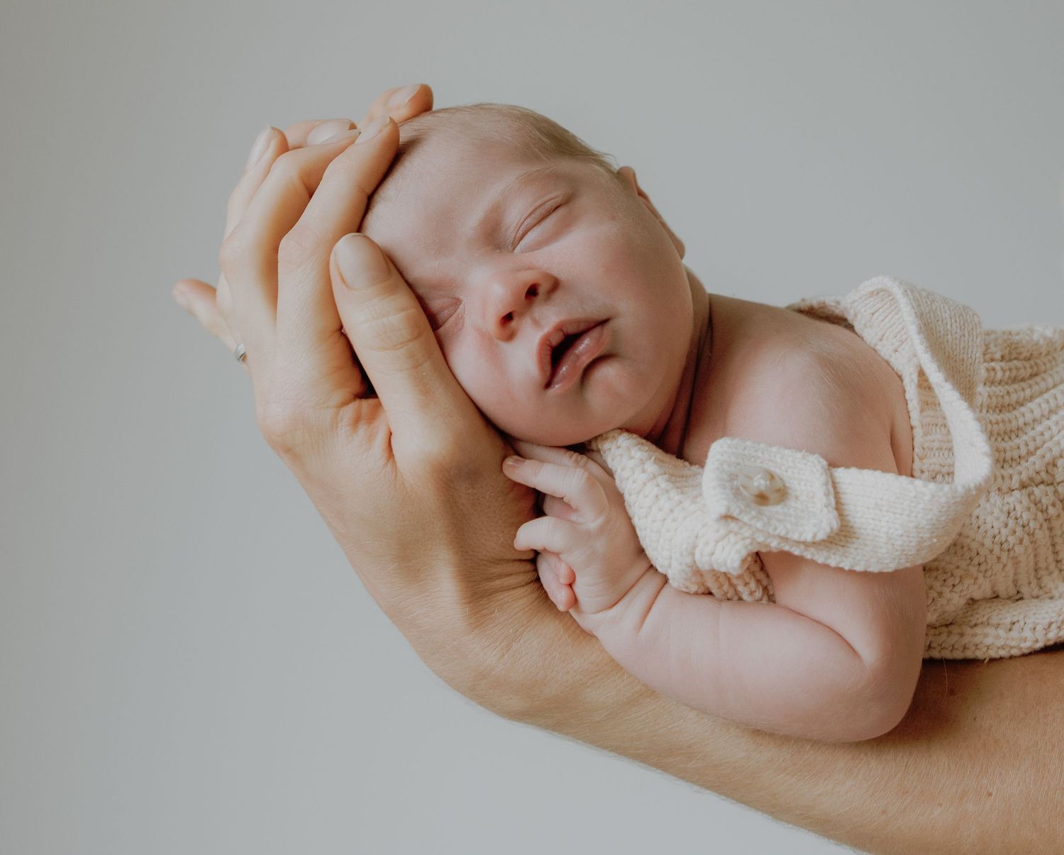 Gentle hands cradle a sleeping newborn baby wearing a soft white knit outfit against a neutral gray background.
