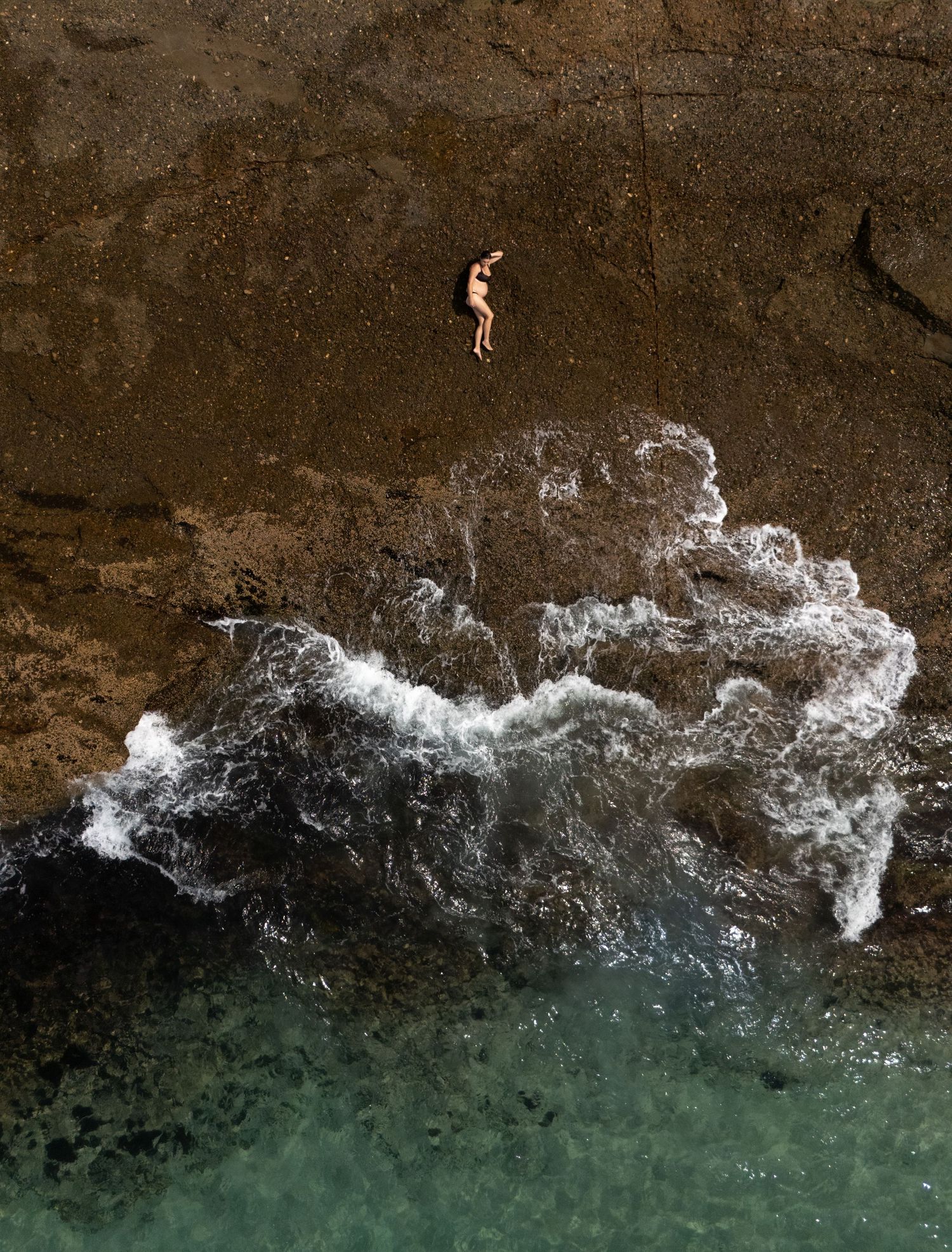 Pregnant mother gently lays on smooth rock while the ocean waves gently pour over the rocks below from the sea.