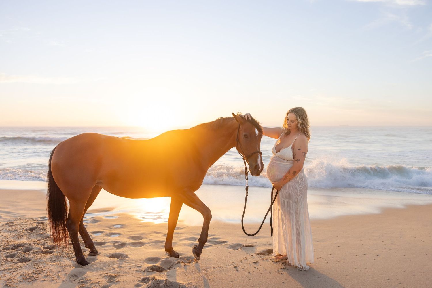 Woman gently strokes horse as the sun rises over the ocean behind them, making the horse glow. The horse is looking at the woman's baby bump