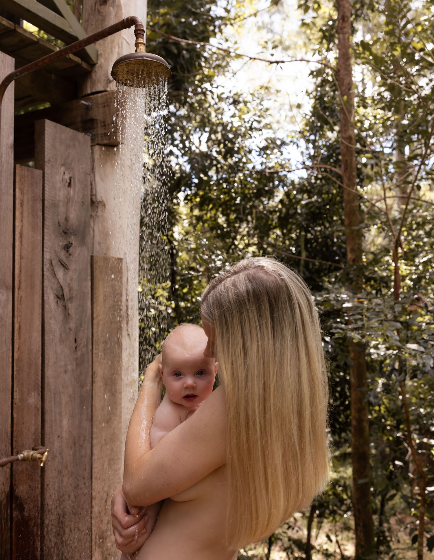 Mother and baby have an outdoor shower with the forest behind them taken at Glenrock. Tap fittings are burnished copper.