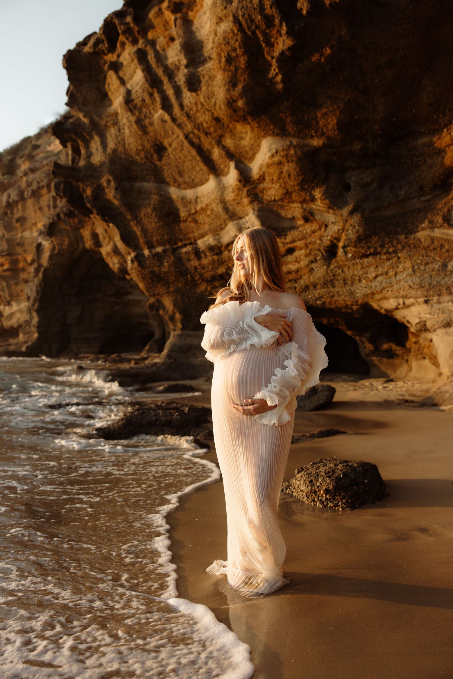 Maternity mum cradling her bump with the soft glow of the morning sun lighting up her face and body while looking out over the water, rocks