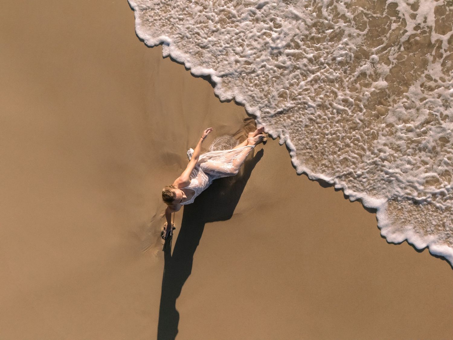 Drone photograph showing pregnant mother laying on her side in the sand at the beach, a small gently foaming wave rolling up near her feet.