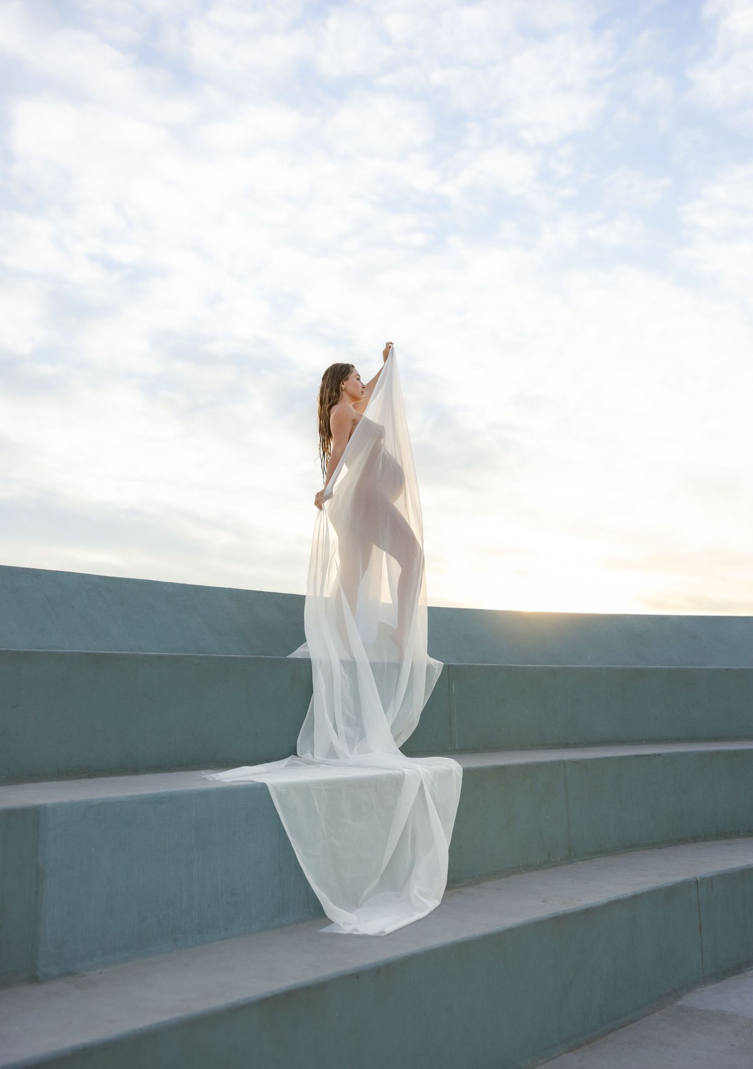 Maternity mum holds white fabric to silhouette her pregnant belly with the sun rising behind the steps.
