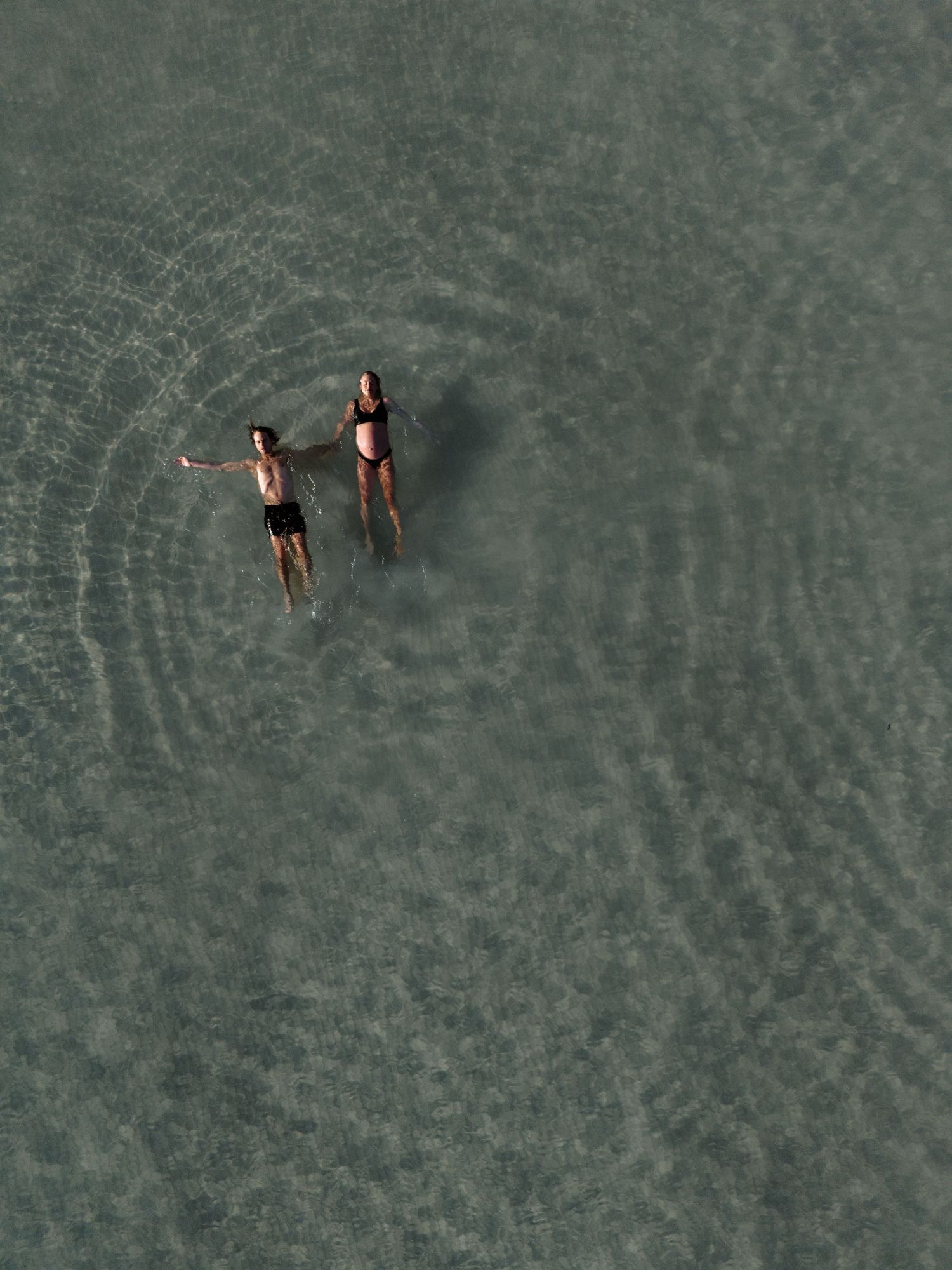 Husband and wife float gently in crystal clear waters of Caves Beach while looking up at the drone.