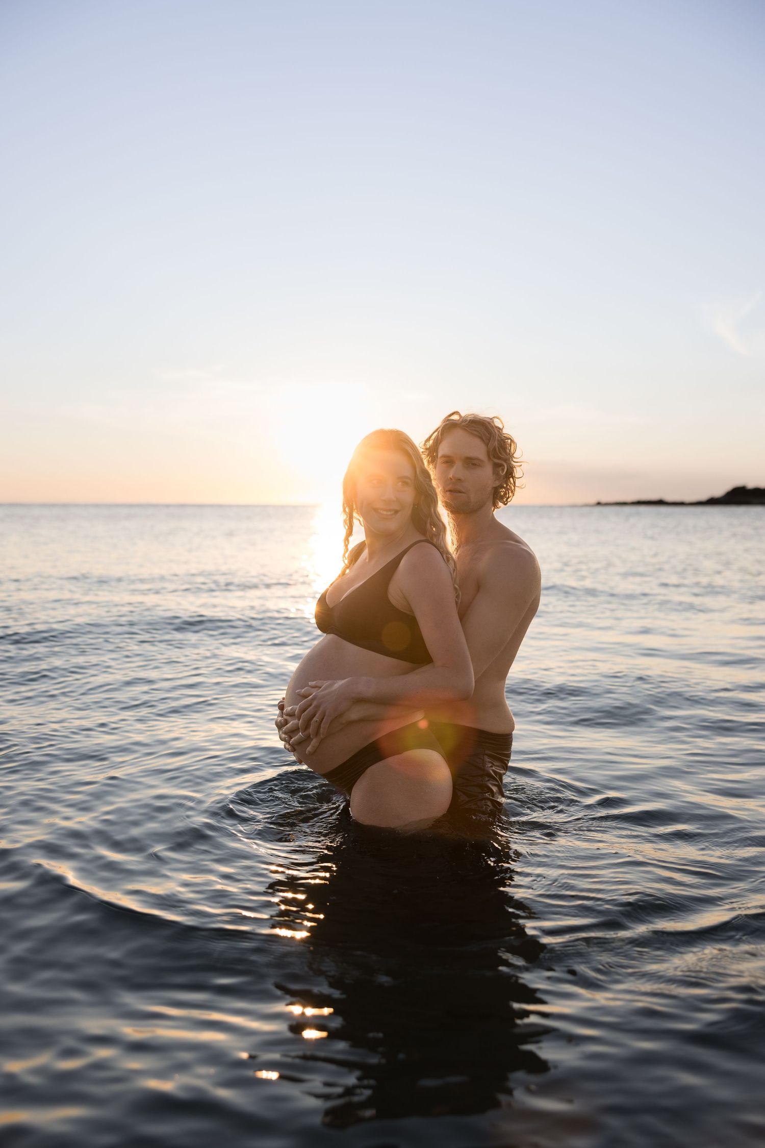 Expecting couple stand knee deep in calm ocean water with the sun rising behind them.