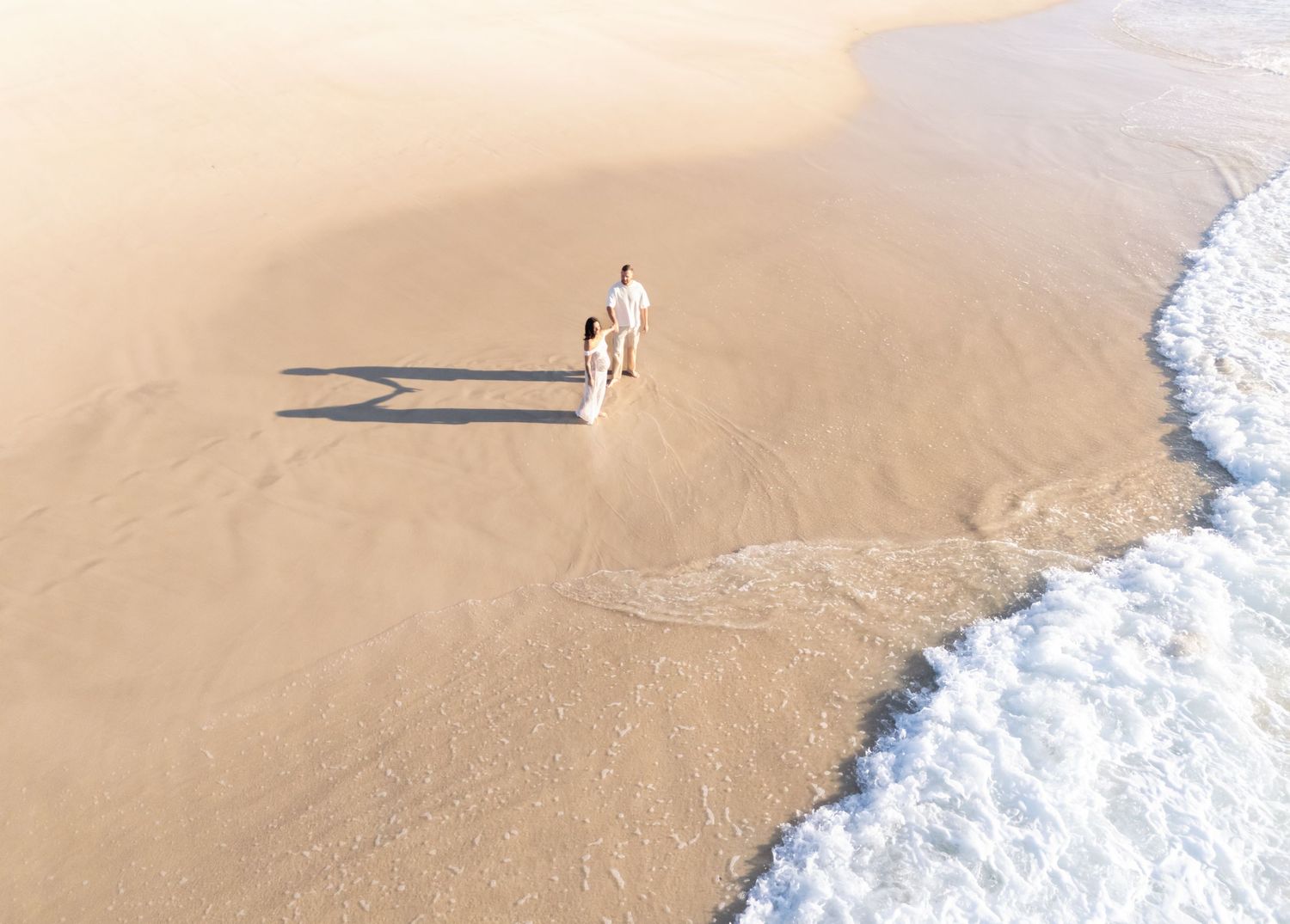 Aerial view of waves gently rolling onto a sandy beach with footprints visible in the wet sand. Husband and wife holding hands.