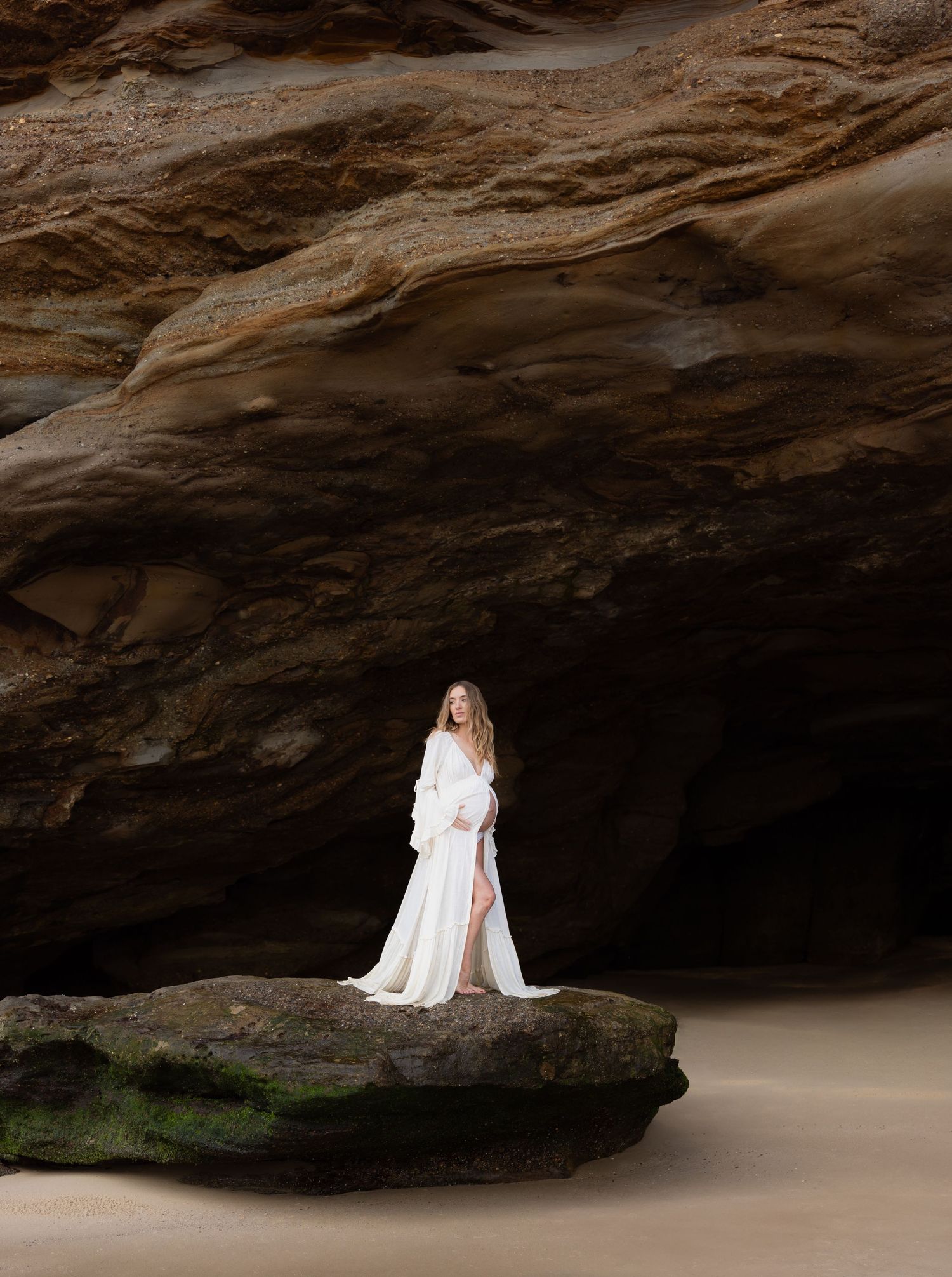 Pregnant woman in a flowing white dress poses gracefully against dramatic rock formations at Caves Beach, Lake Macquarie.