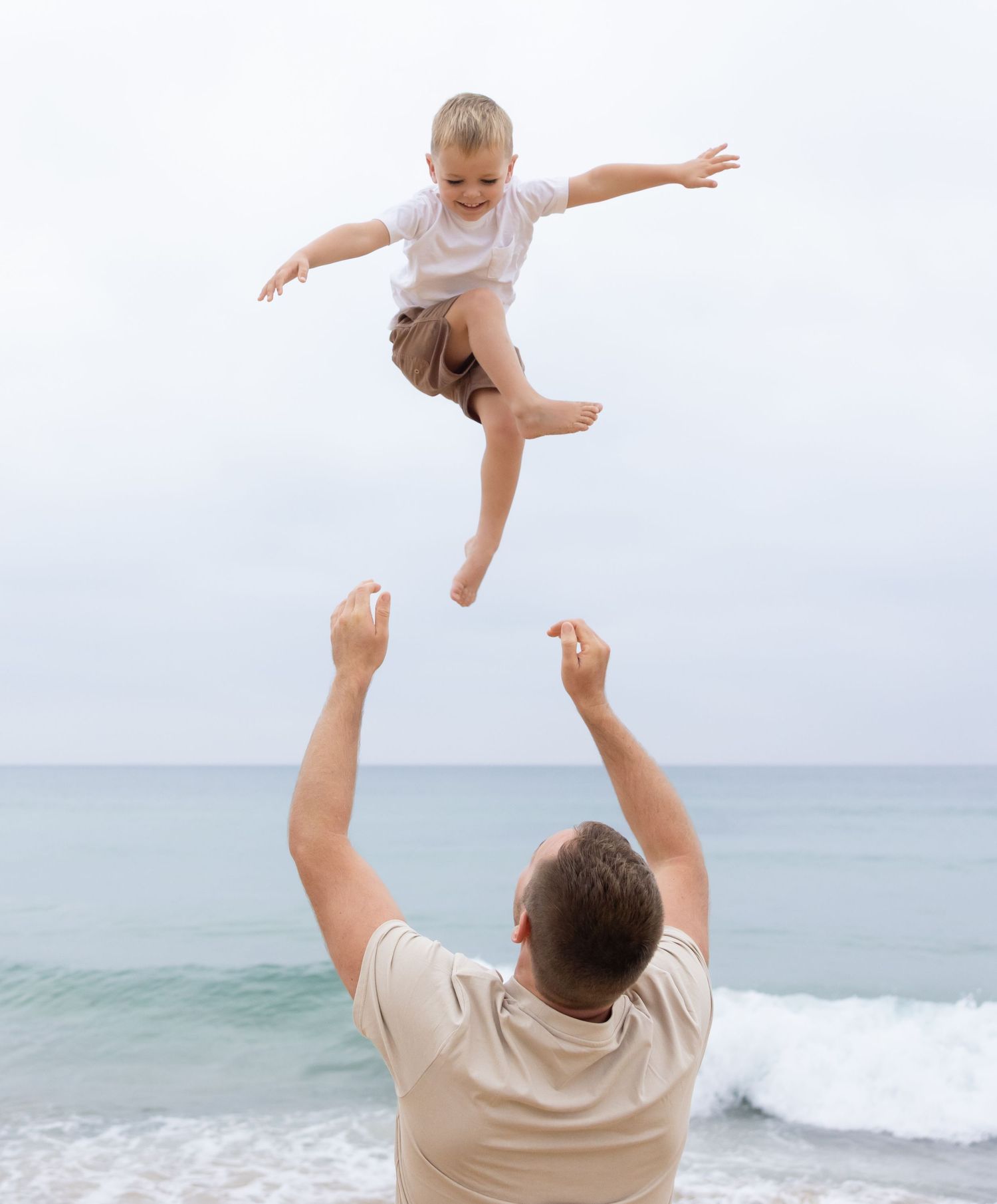 Father throws boy into the air. Boy performs a super hero pose mid throw beaming.