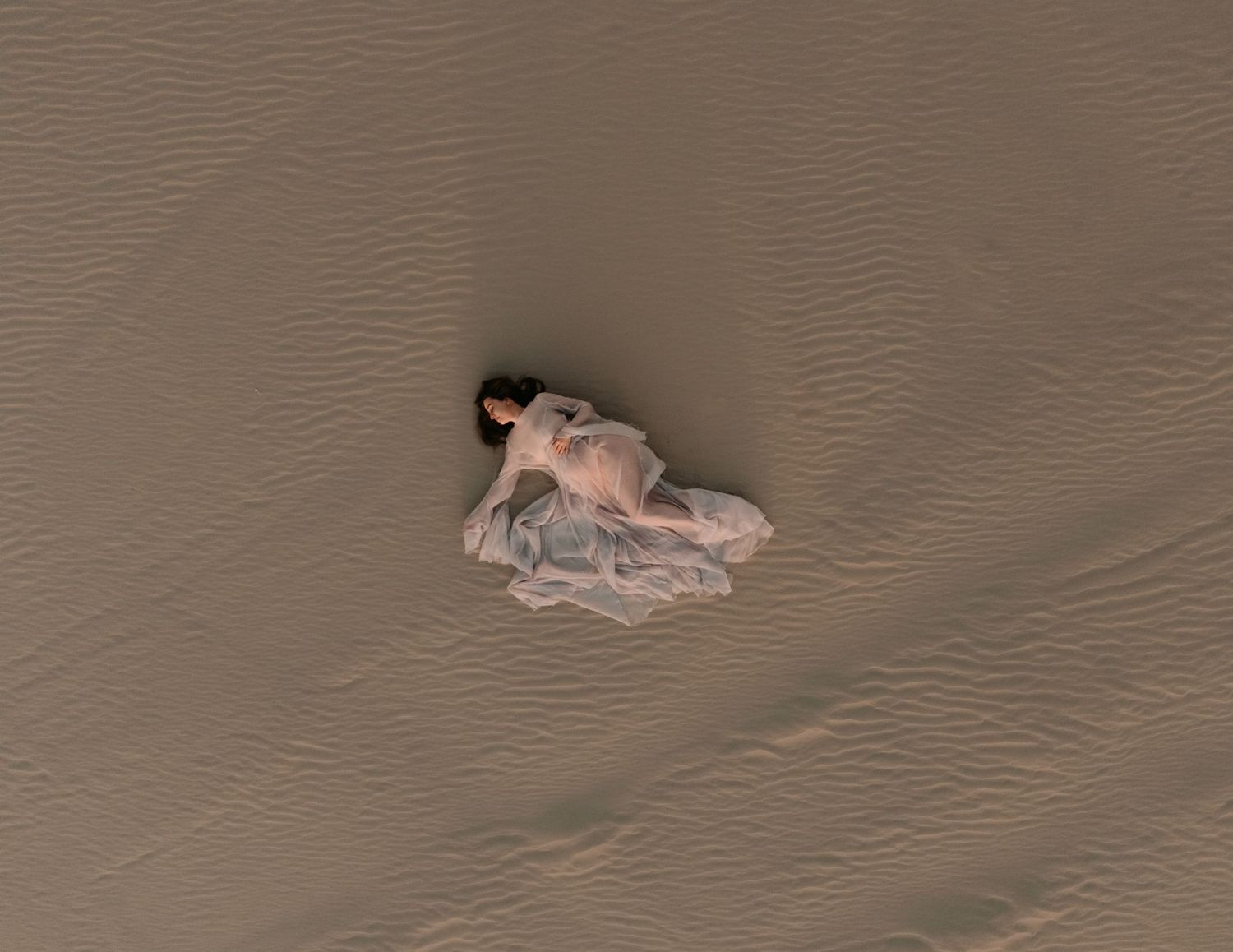Aerial image of pregnant mum laying in the rippled sand of Port Stephens sand dunes in a billowing  cream dress.