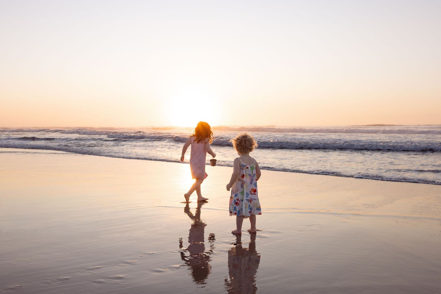 Sisters run along the shallow waters at Bar Beach as the sun rises over the ocean. They are playing together with a bucket and spade.