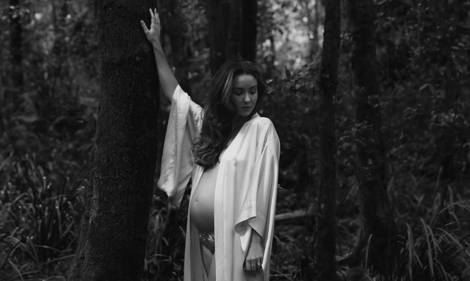 Pregnant mother in robe leans against a tree in the forest at Glenrock lagoon in Newcastle.