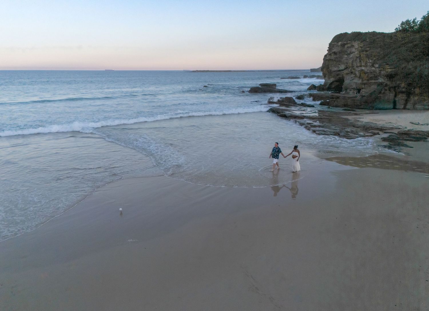 Drone image of man and woman walking hand in hand at sunrise at Caves Beach.