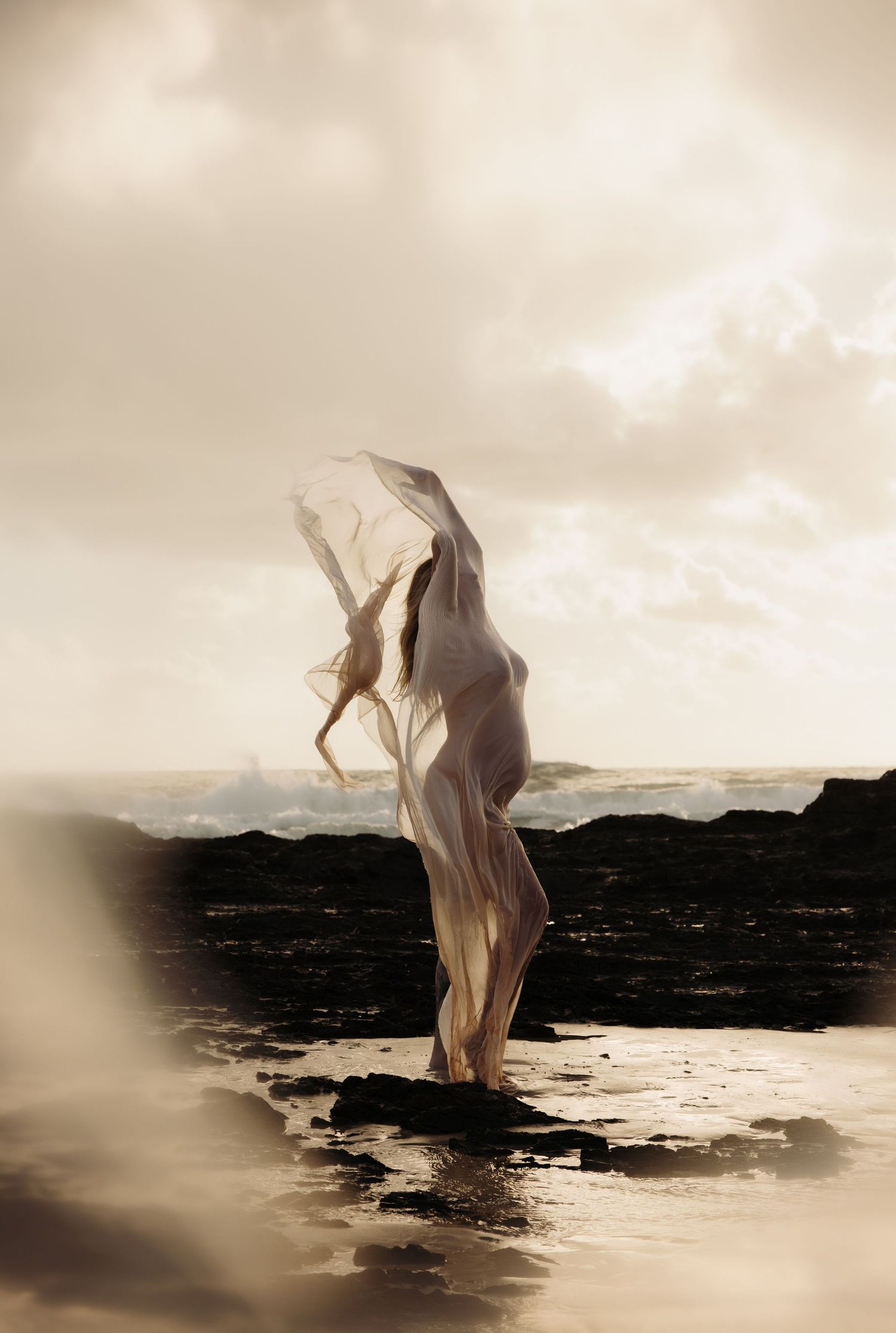 Ethereal figure in flowing white fabric dances along a moody rocky coastline at dusk, her pregnant belly curved under the fabric.