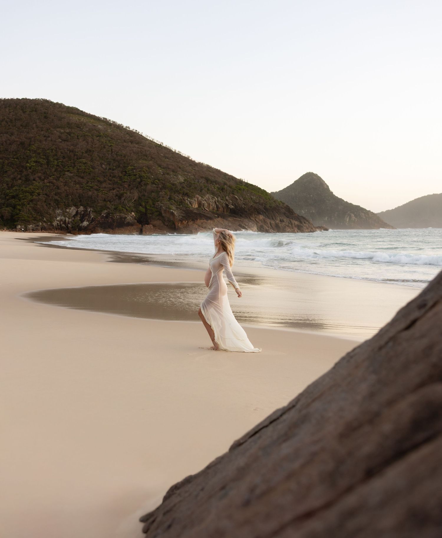 Pregnant mum at sunrise with the wind in her hair and dress flowing behind her. The ocean and coastal headlands are pictured behind her.