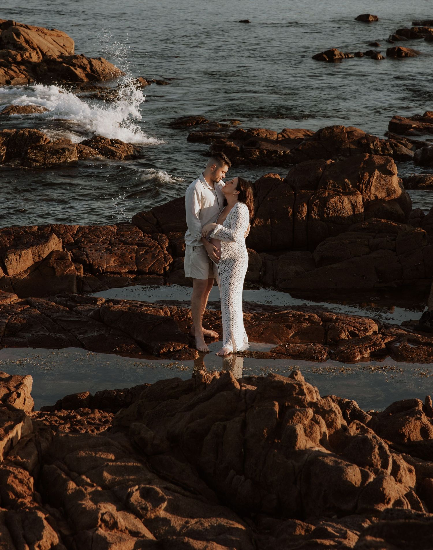Couple adventuring over the rocks, waves splashing behind them, ready to embrace.