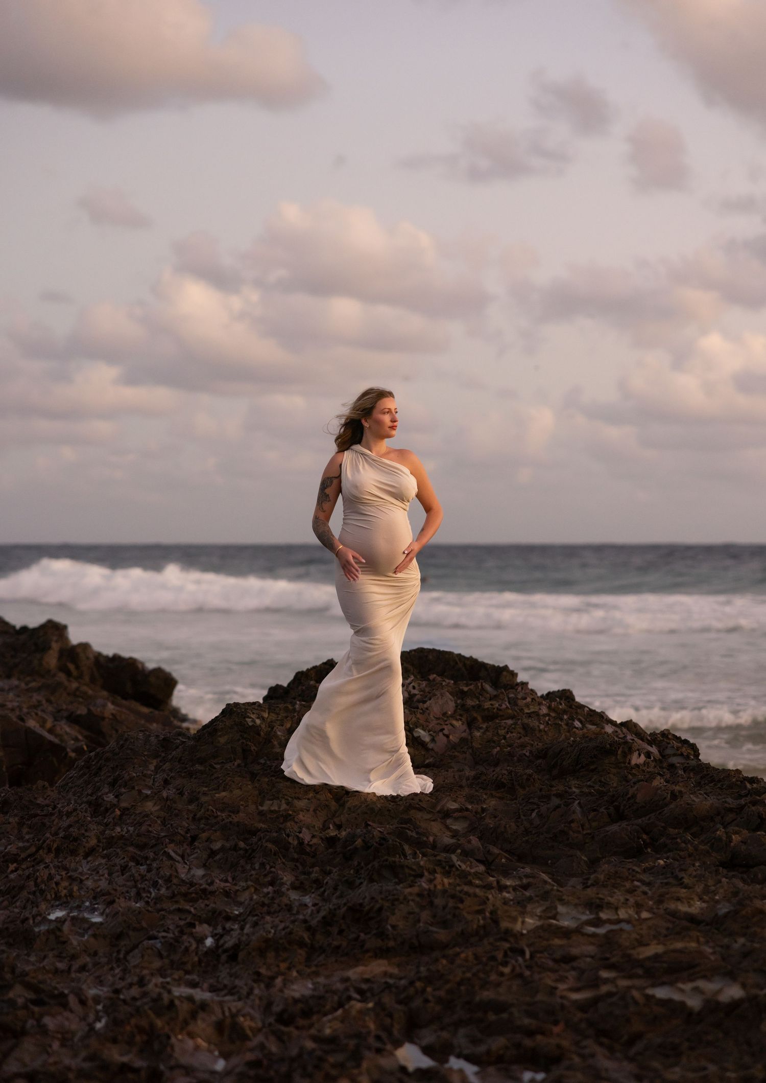 A figure in a flowing white dress poses dramatically on dark rocks against a moody ocean backdrop at sunset.