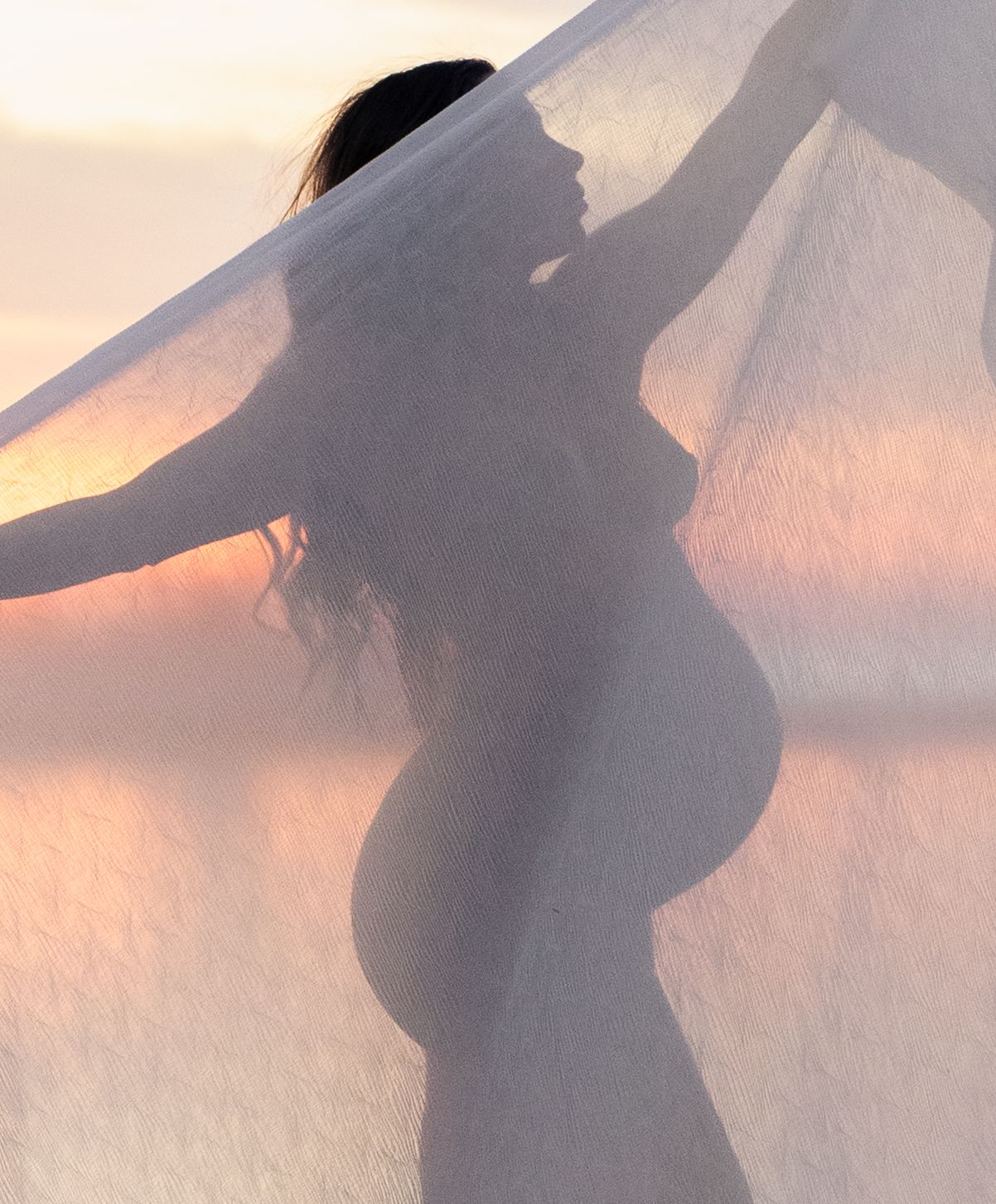Silhouette of pregnant mother behind fabric at sunrise.