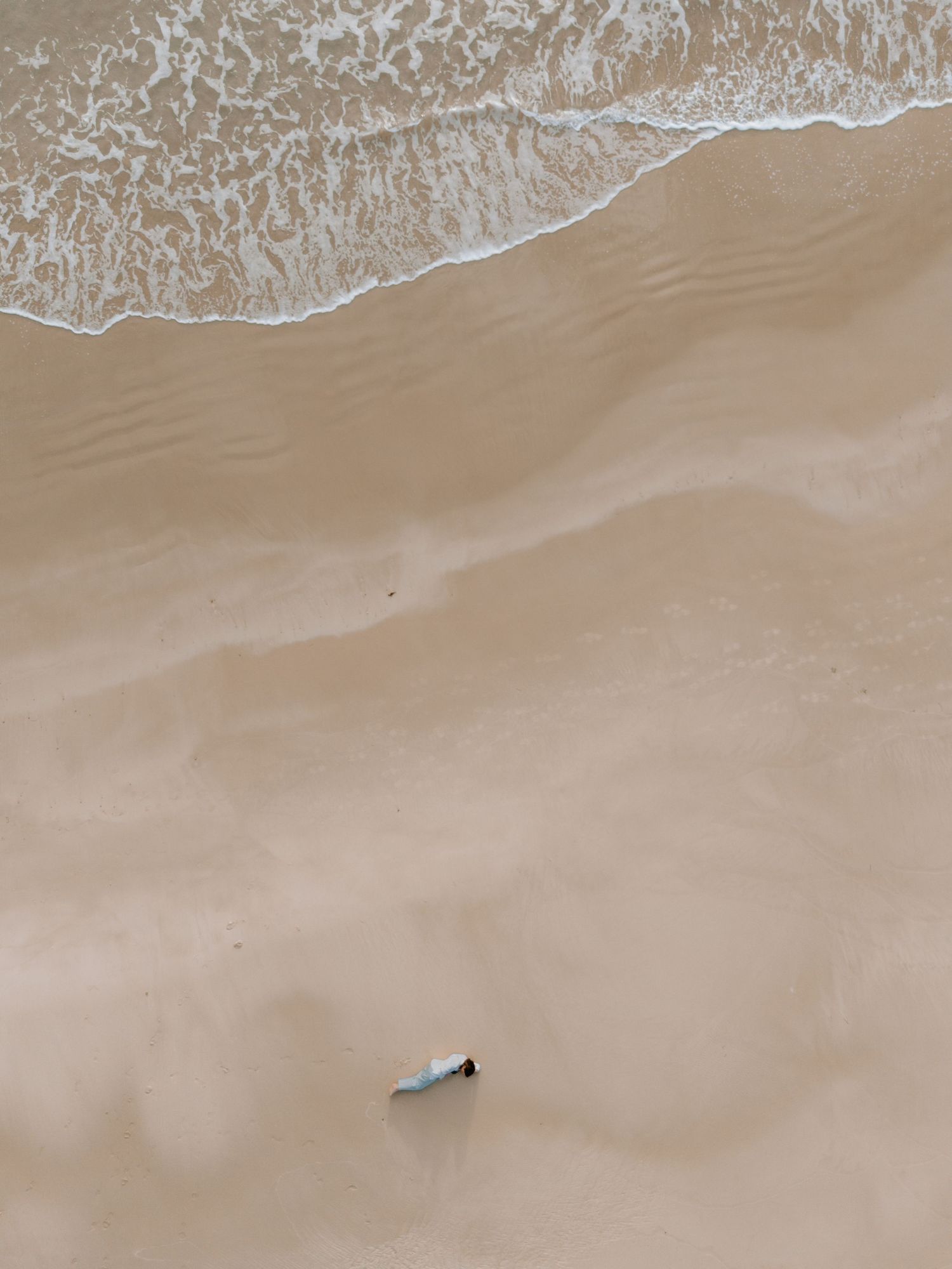 Woman lays on beach with ocean waves rolling up below her.
