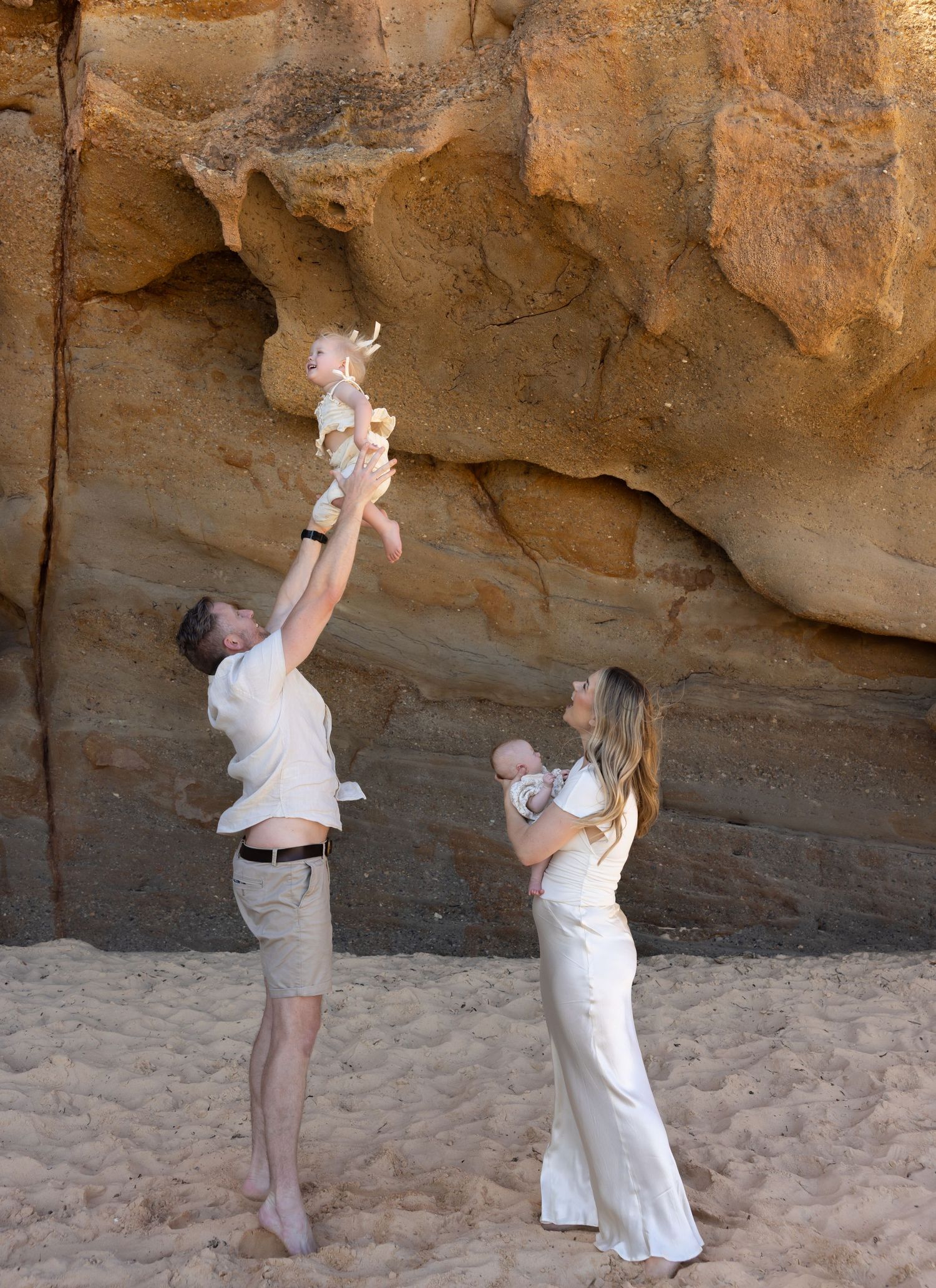 Family moment captured father plays with oldest daughter, gently reaching her into the air. Mum holds baby sister and looks up at daughter