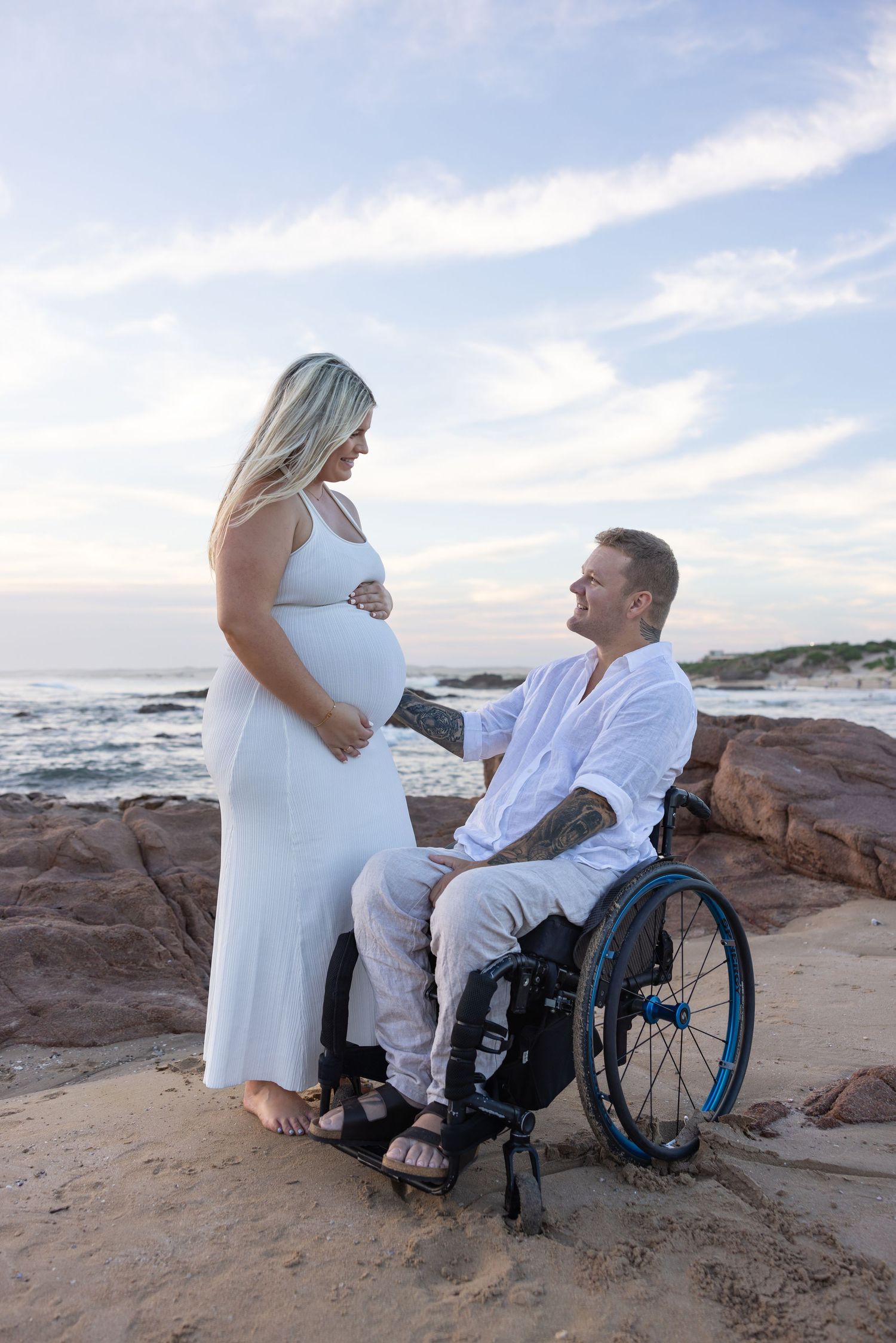 Pregnant mum standing next to her partner who is in a wheel chair looking at each other lovingly. Ocean stretches behind and clouds in sky.