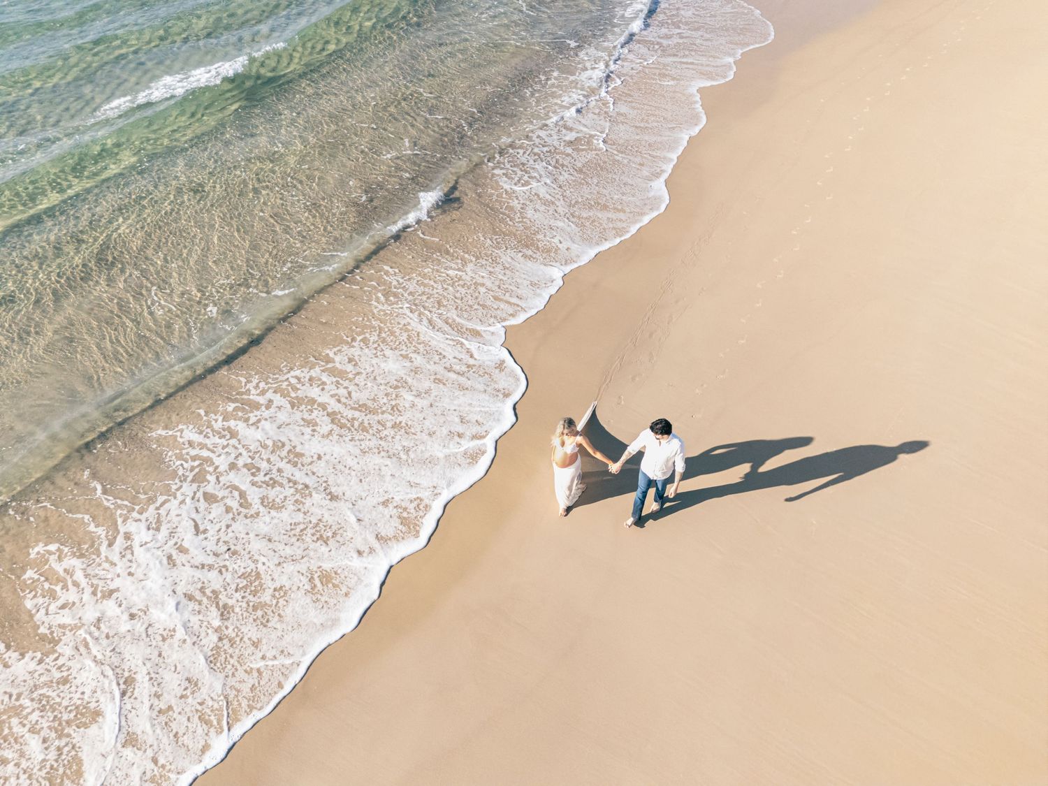 Lady and man stroll along the beach at sunrise, their shadows casting a love heart shape behind them, captured by drone.