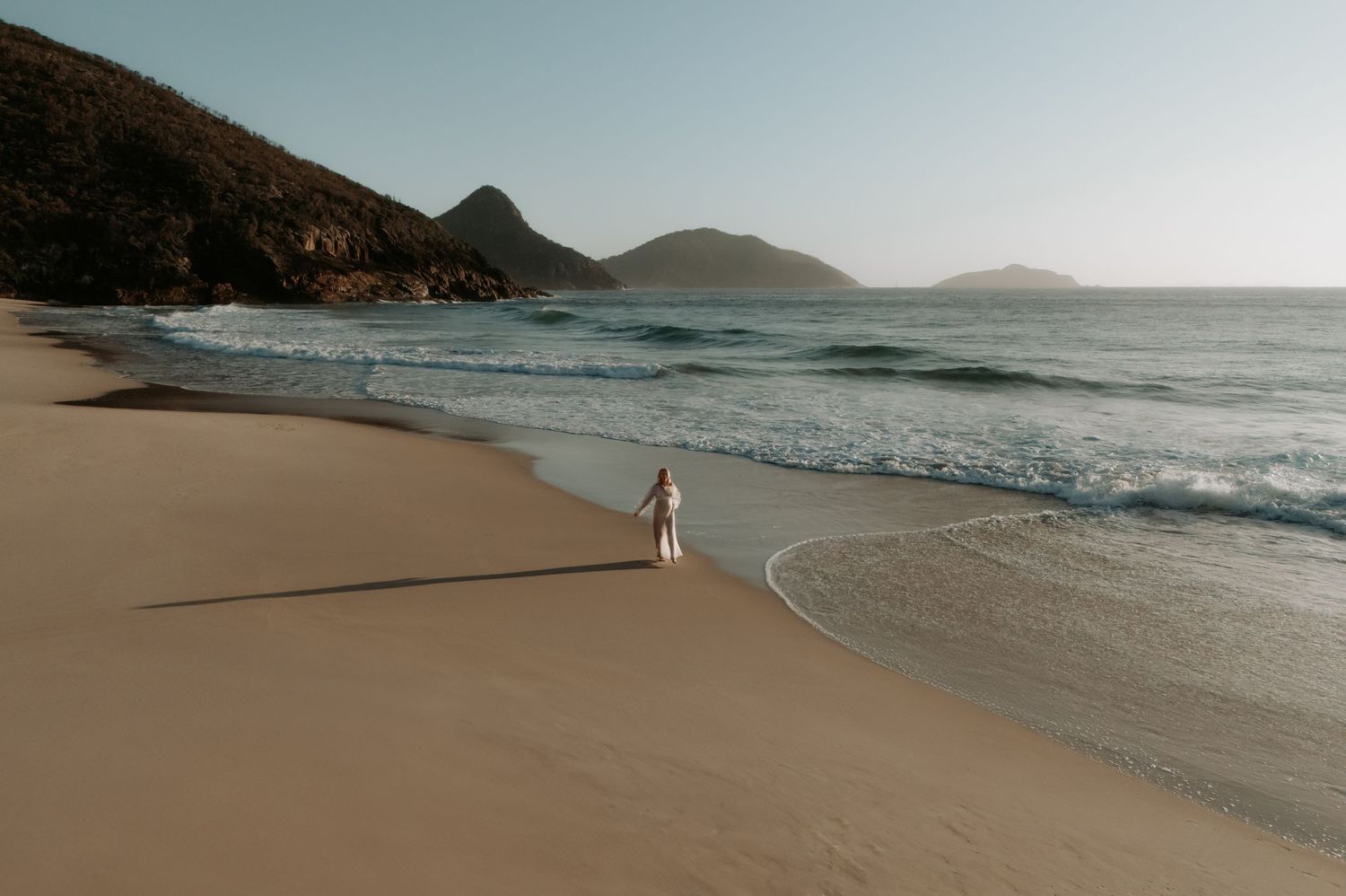 Pregnant mum framed by drone in a white dress with the ocean and Tomaree Headland in the distance.