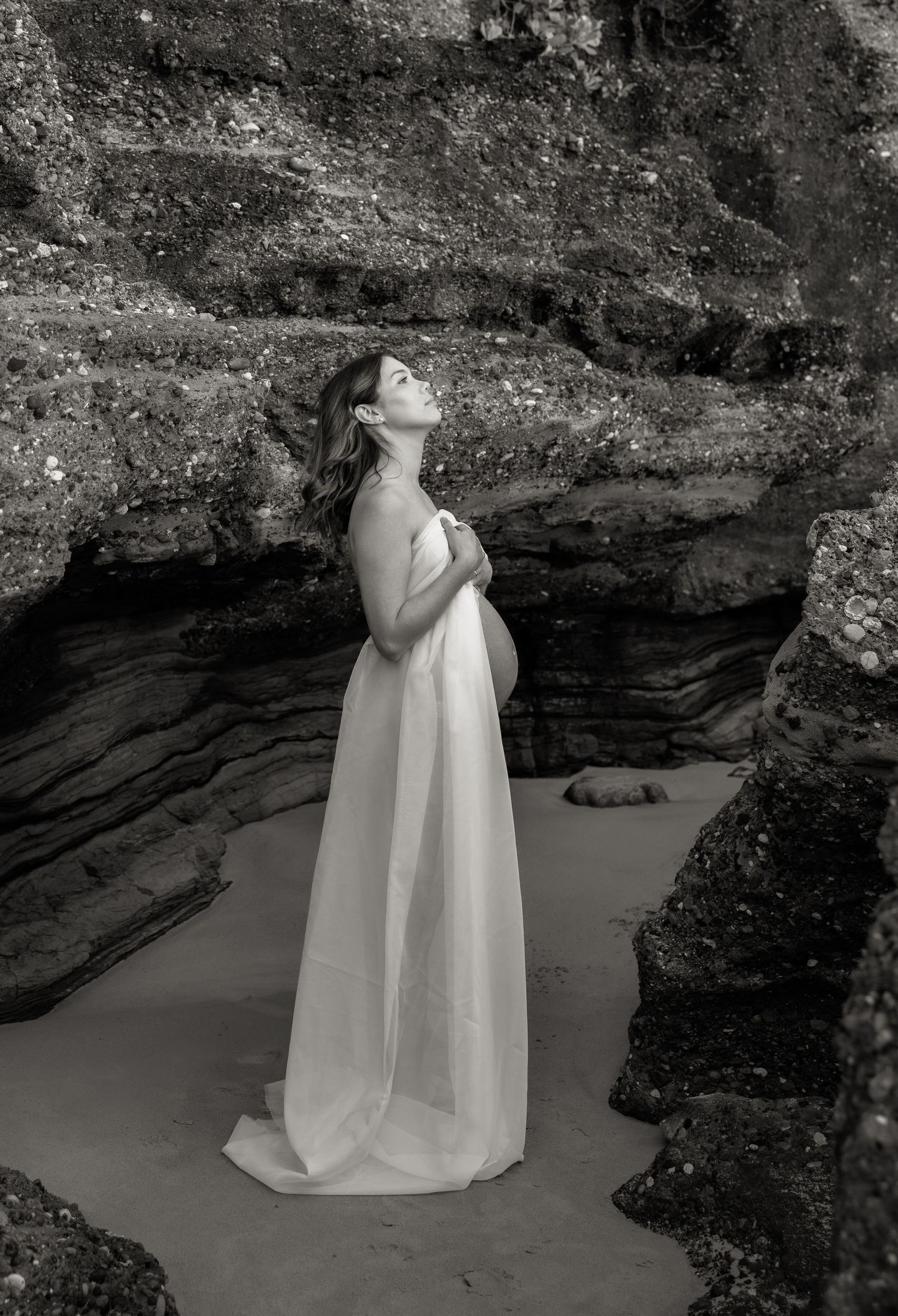 Pregnant mum stands with fabric wrapped around her, belly showing, rocks behind in black and white.