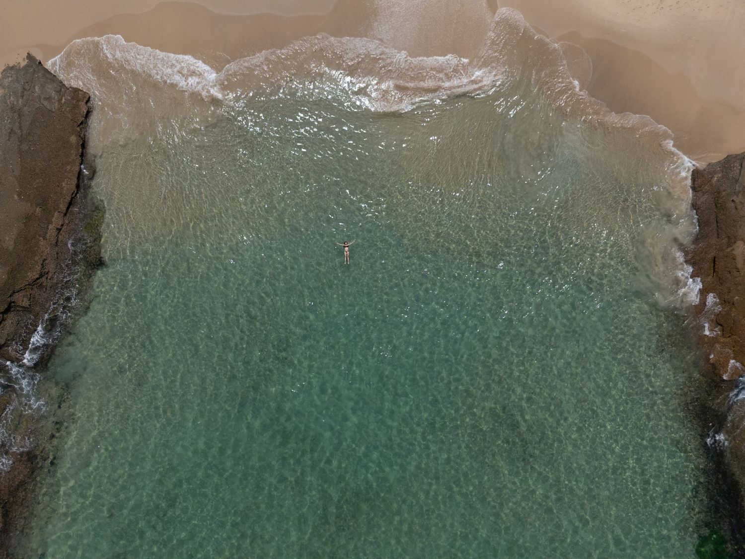 Aerial view of turquoise ocean waves crashing against a sandy beach shoreline creating white foam patterns.