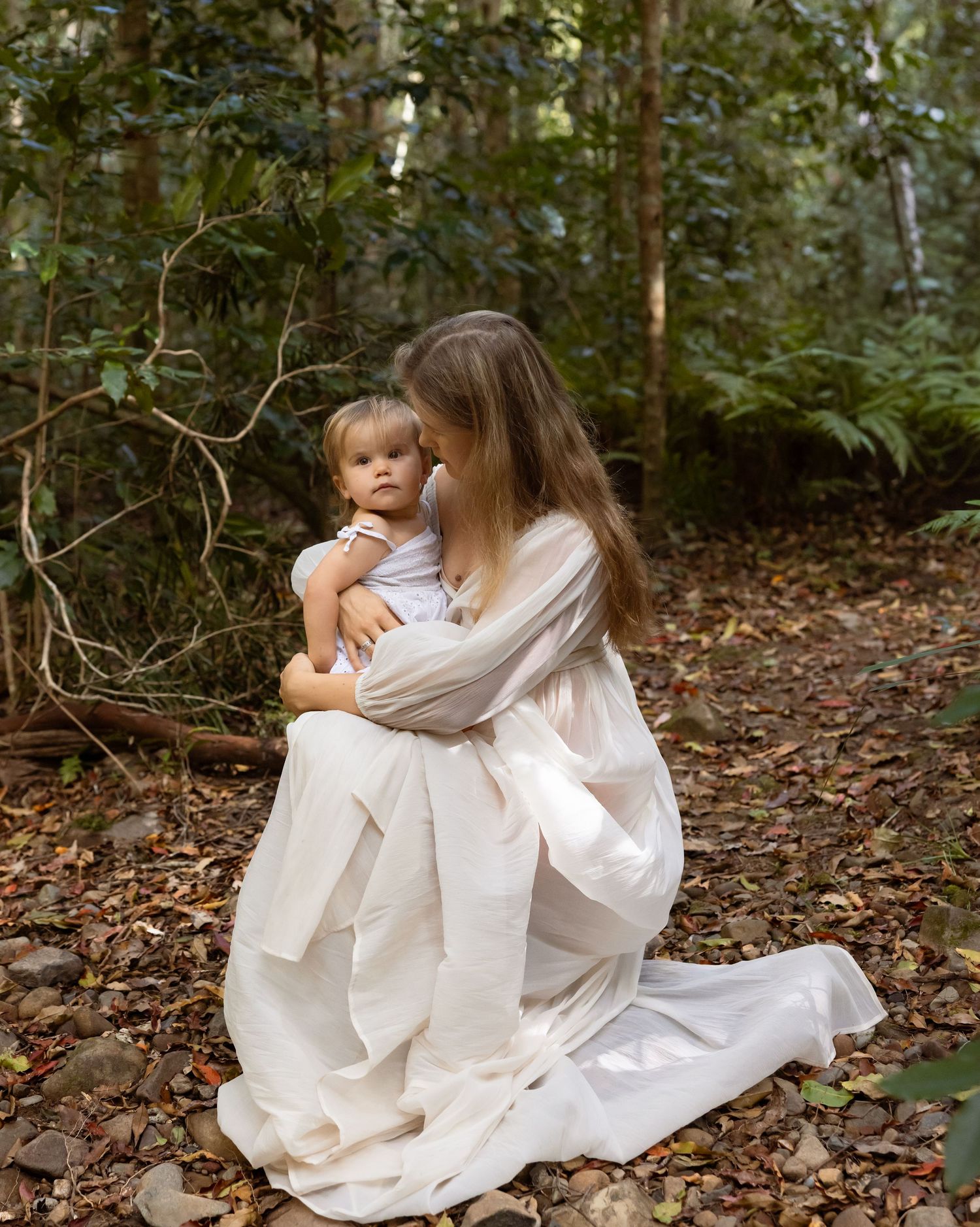 Mother holds daughter on her knees as she bends low on the tree leave covered forest floor.