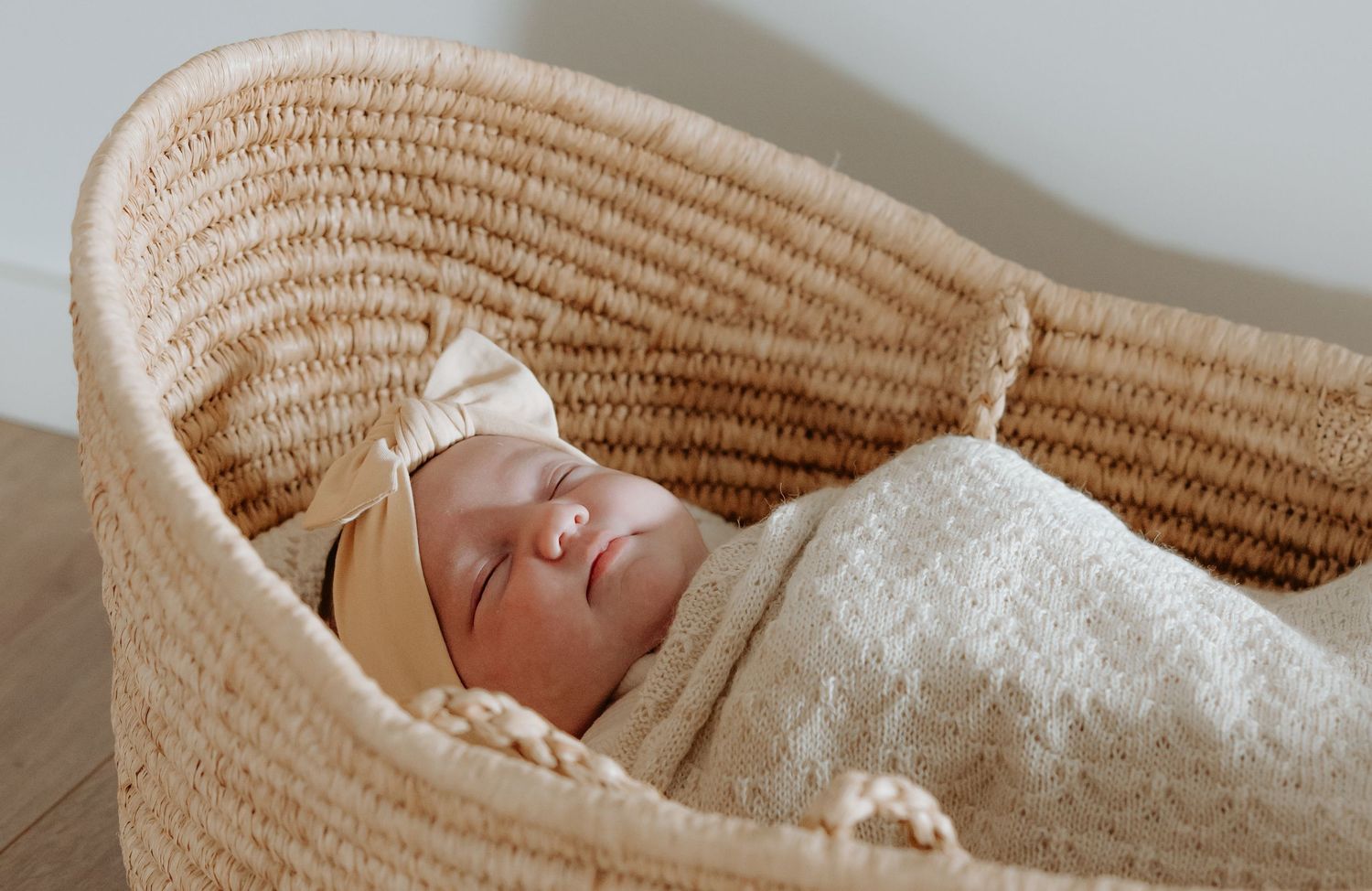 Newborn baby laying in a seagrass baby basket, fast asleep wrapped in a wool blanket with a bow in her hair.