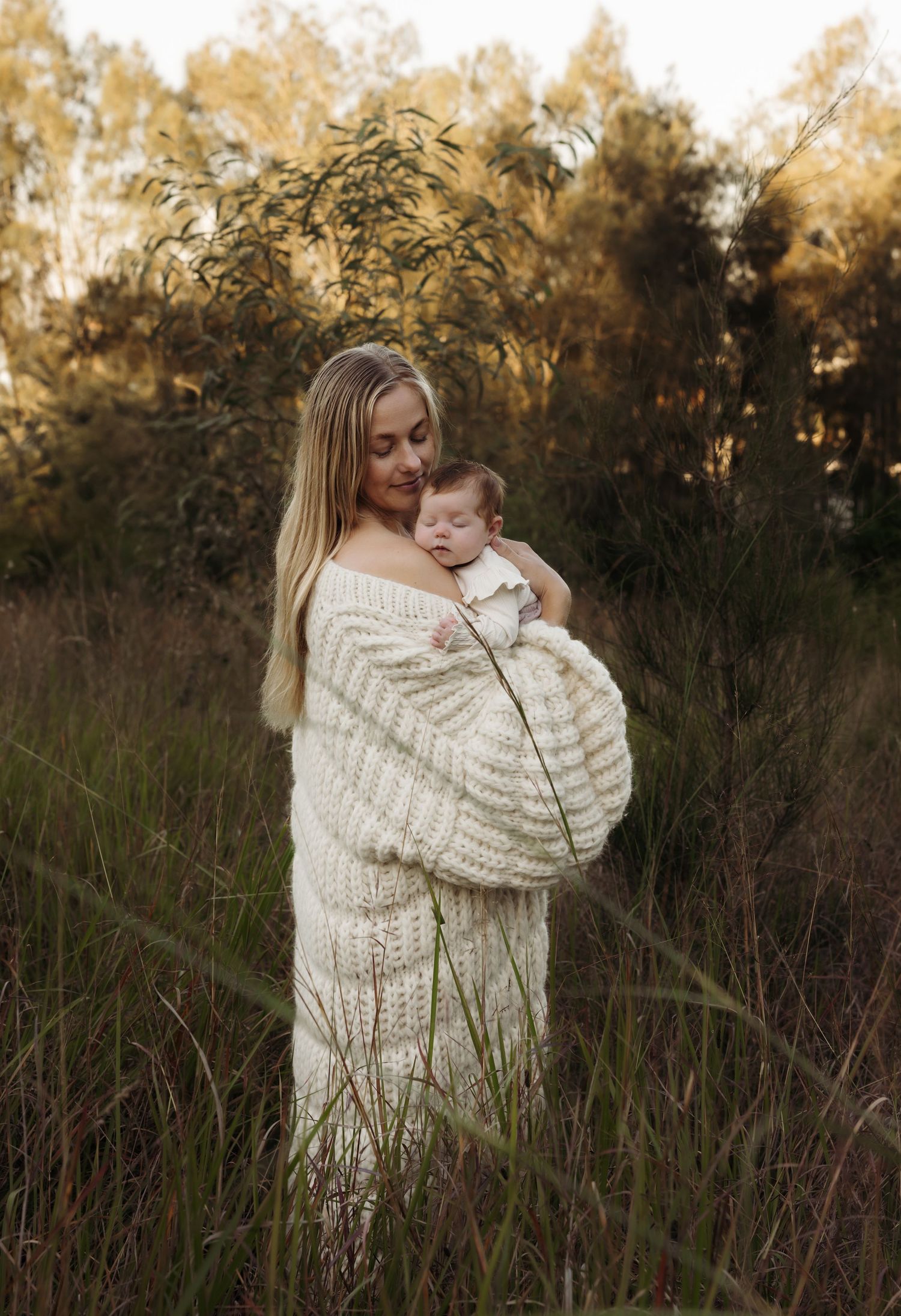 Mother holding baby daughter amongst the tall brown grass field at Booragul with trees in the background as the sun sets casting shadows.