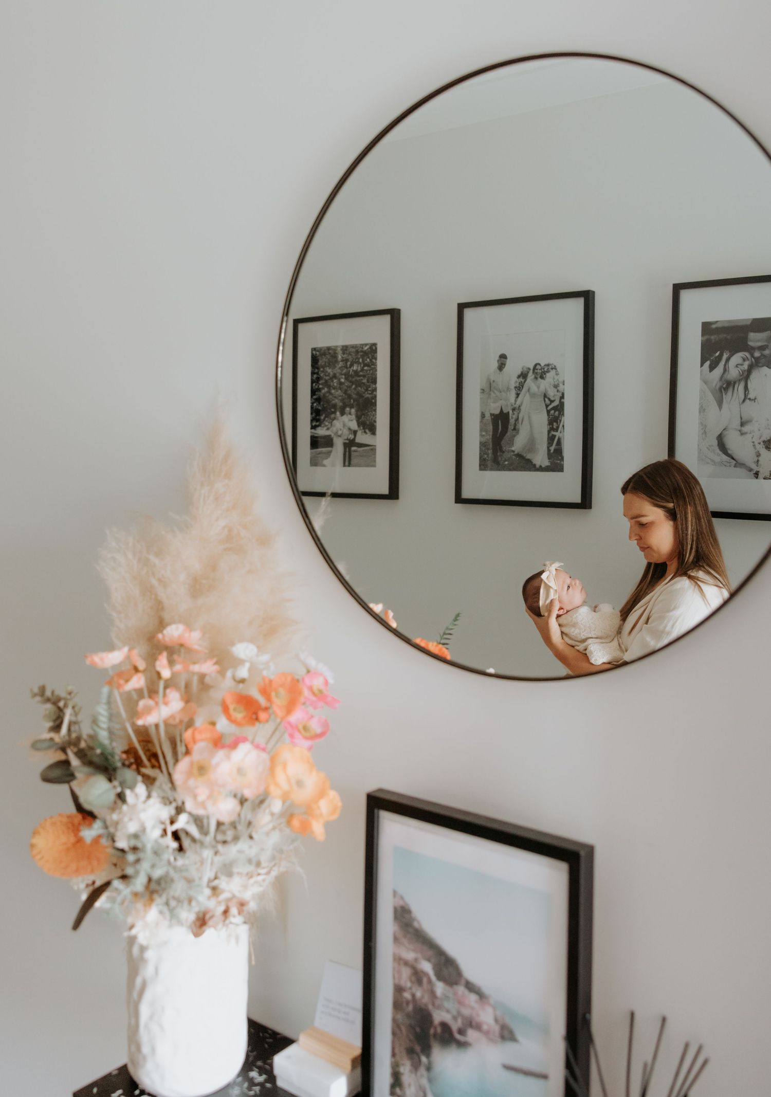 Mother and baby daughter look at each other. Photo taken through hallway mirror with flowers on the side table and photographs on the walls.